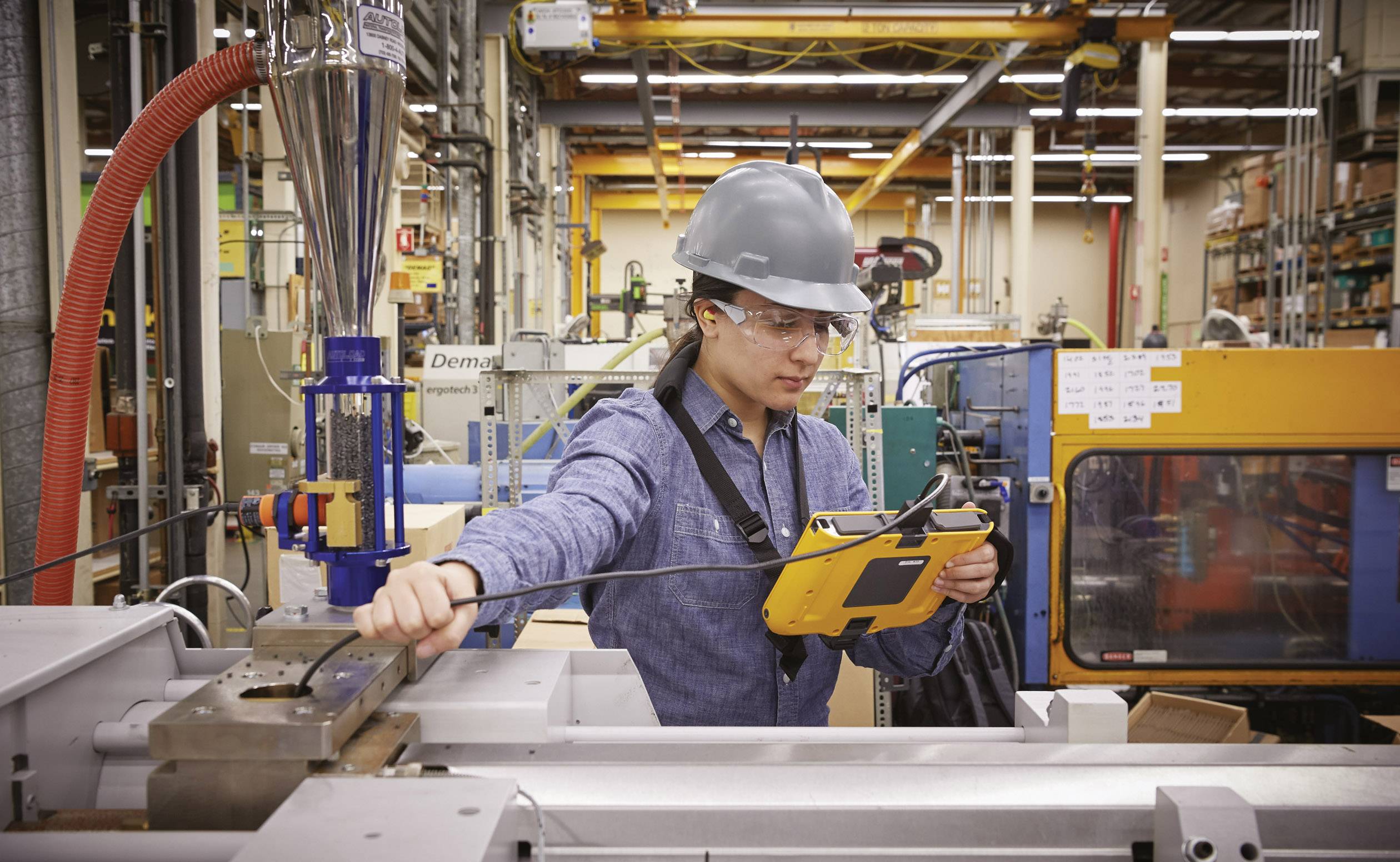 Een man met een veiligheidshelm bedient een machine in een fabriek. Hij houdt een tablet in zijn hand en staat omringd door technische apparaten.