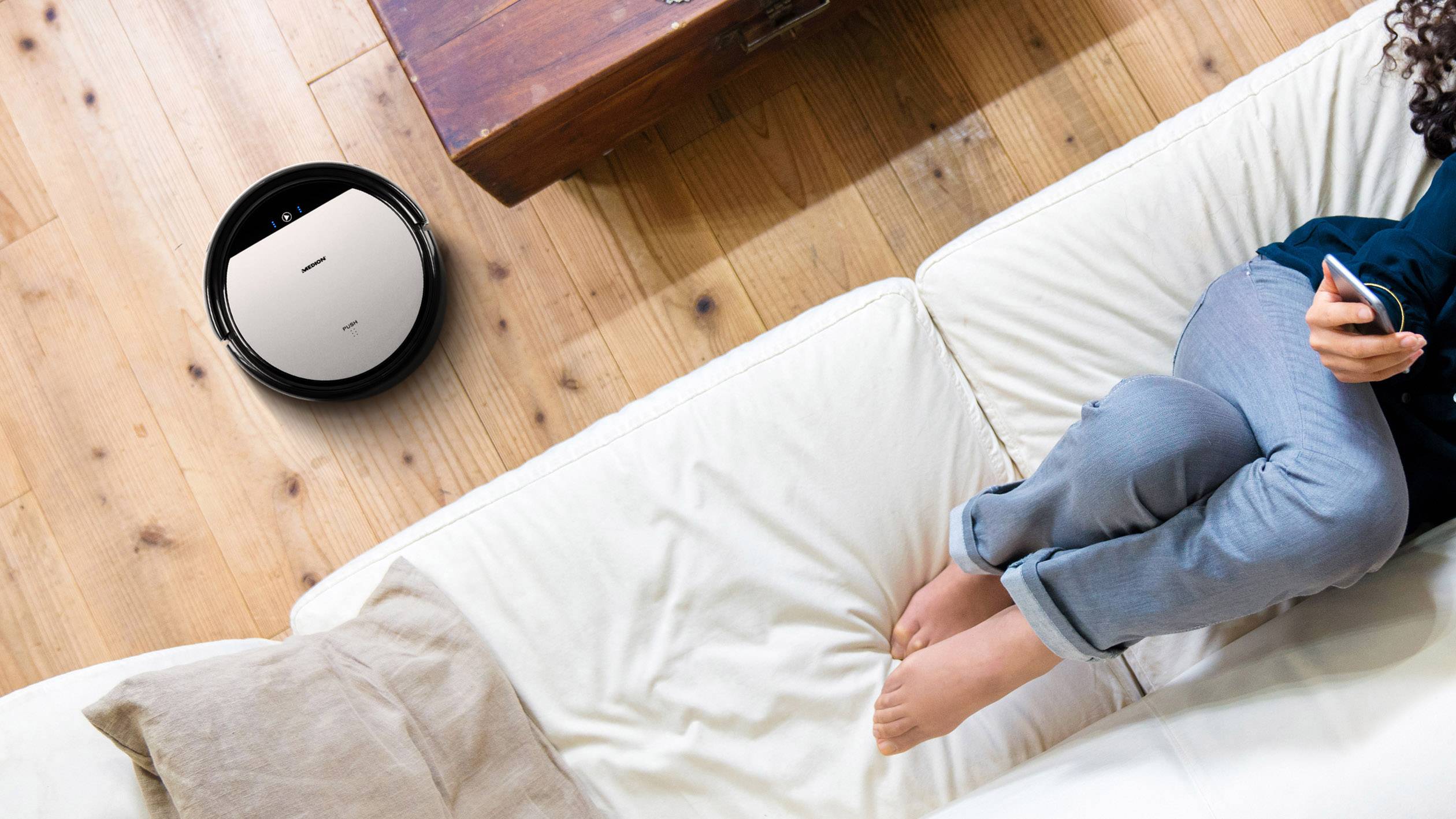 Een persoon zit op een bank en houdt een smartphone vast terwijl een ronde robotstofzuiger de houten vloer schoonmaakt.