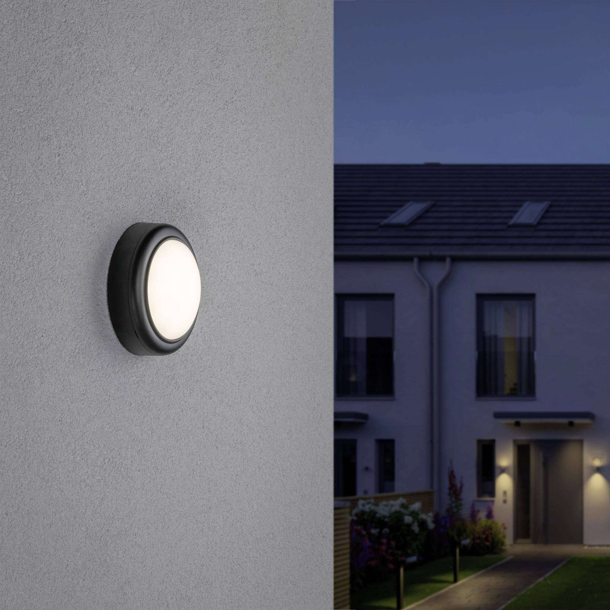Buitenlamp aan grijze muur verlicht binnenplaats met moderne gebouwen op de achtergrond bij schemering.