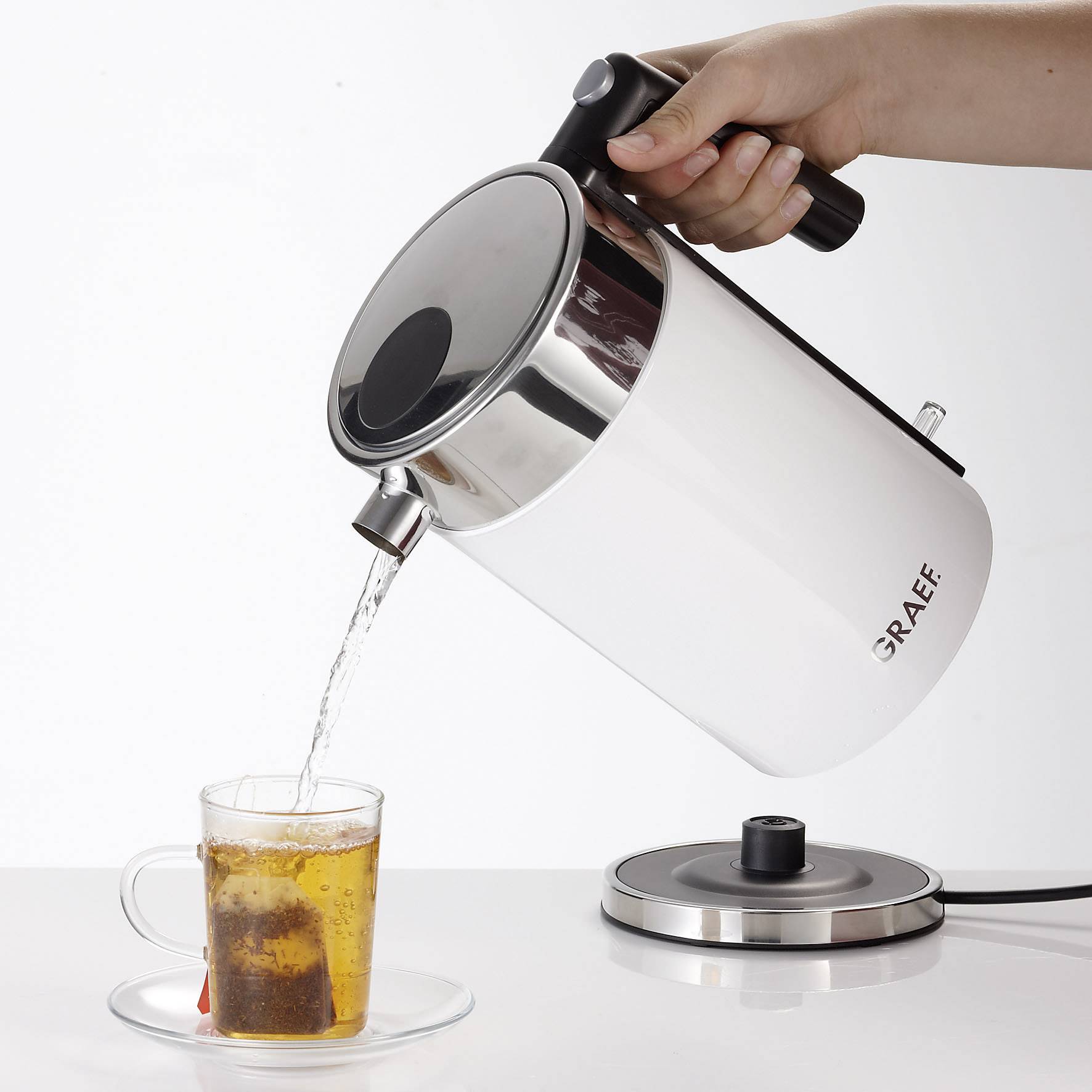 Een hand giet water uit een elektrische waterkoker in een theekopje. De waterkoker staat op een basis en is voorzien van het 'Graef' label.