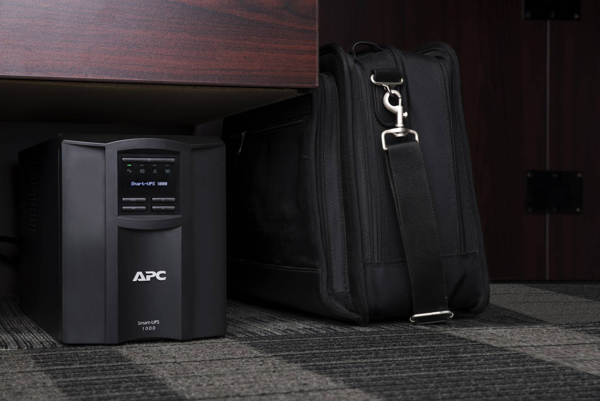 Een APC-UPS-apparaat dat onder een tafel staat naast een zwarte aktetas op een tapijtvloer.