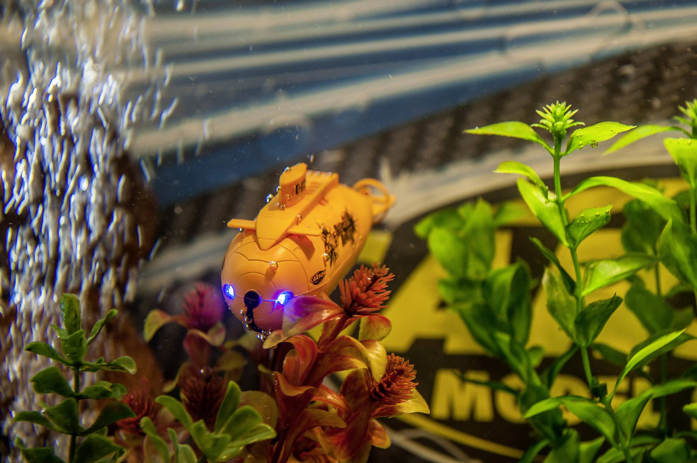 Een kleine oranje speelgoed-onderzeeër met blauwe lichten drijft in een aquarium, omgeven door planten en luchtbellen.