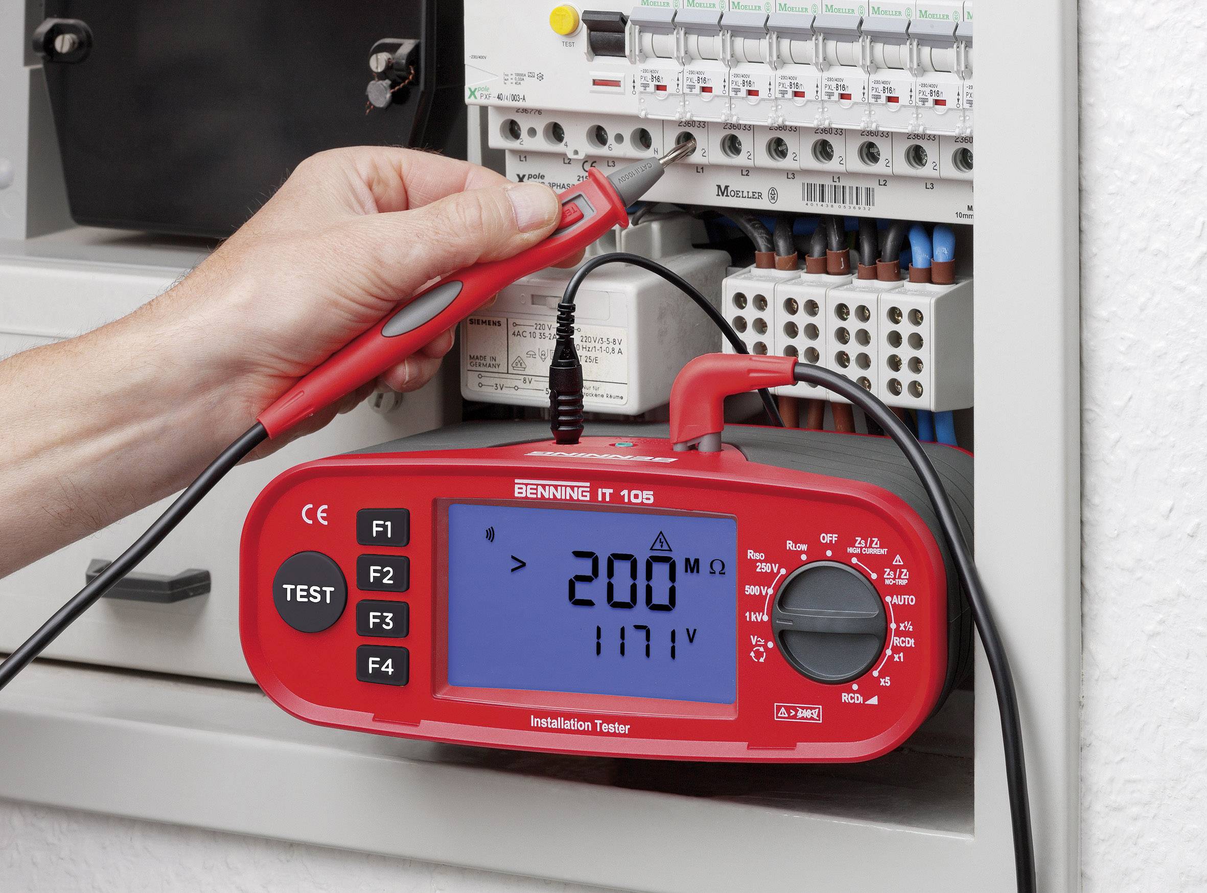 Een persoon gebruikt een rood installatiemeetapparaat om de spanning te meten in een open zekeringkast. Het display toont '200V' en '117V'.