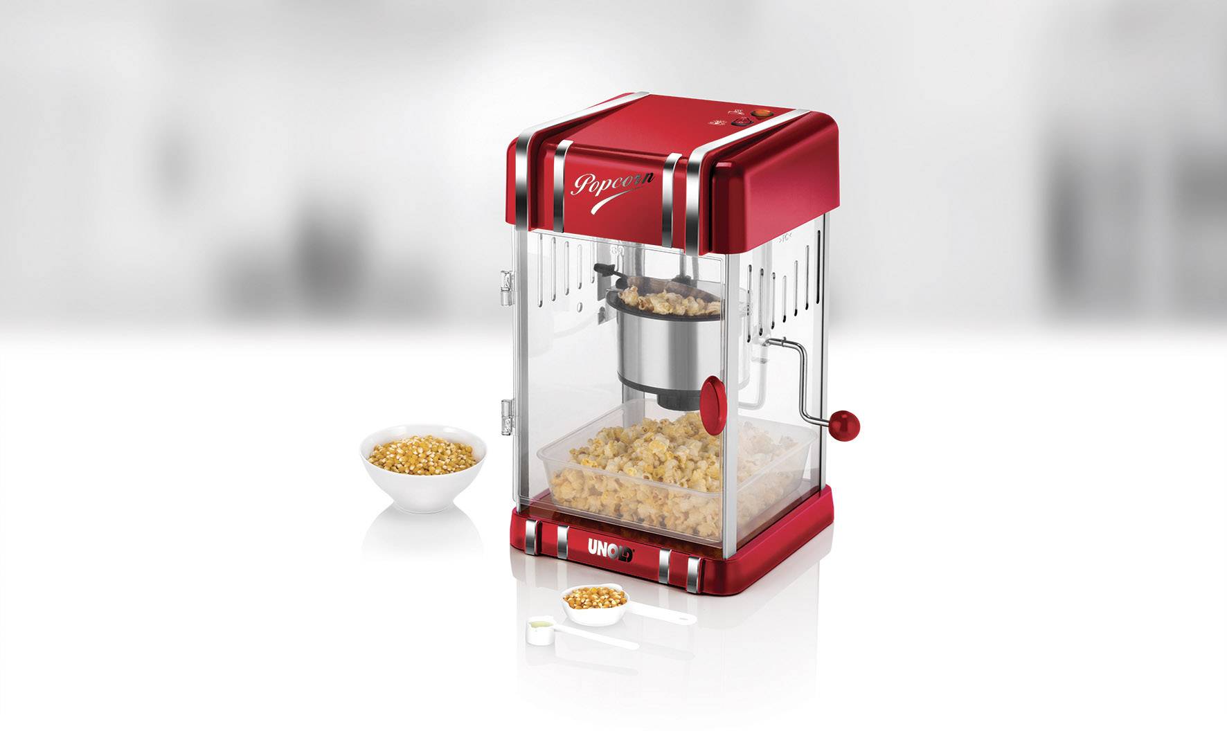 Rode popcornmachine met glazen behuizing, naast een kom met maïskorrels en een maatlepel. De machine staat op een wit oppervlak.