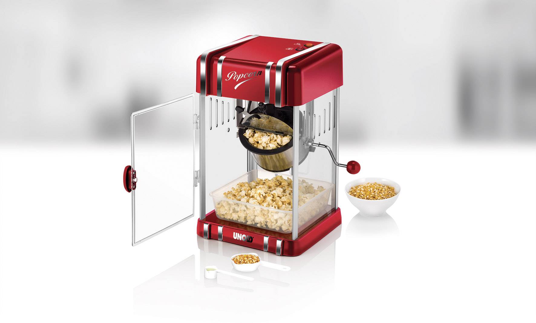 Rode popcornmachine met open deurtje, gevulde kom aan de binnenkant. Ernaast staat een kom met maïskorrels en een maatlepel.