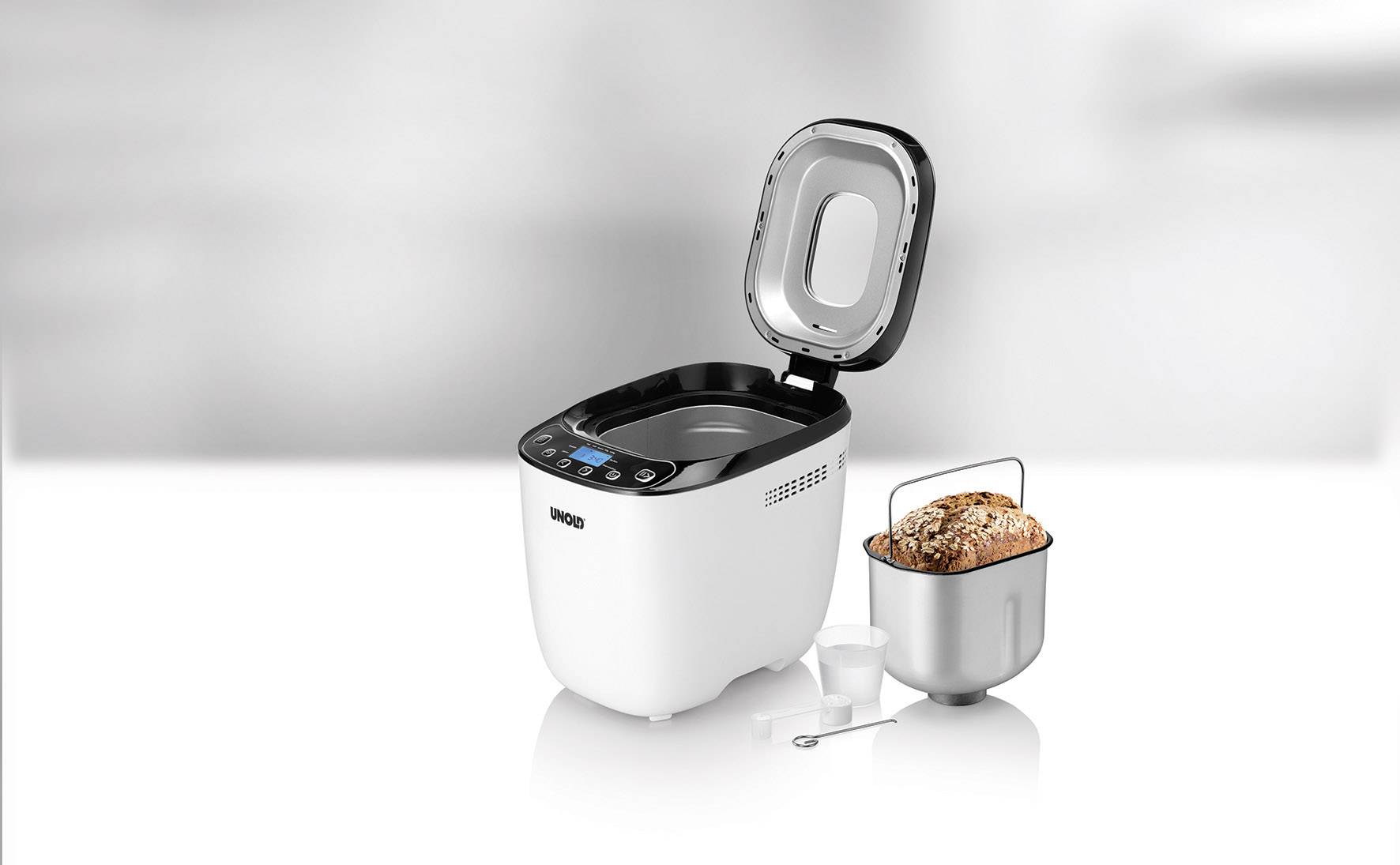 Broodbakmachine met open deksel, broodvorm met gebakken brood ernaast, maatlepel en kleine maatbeker.