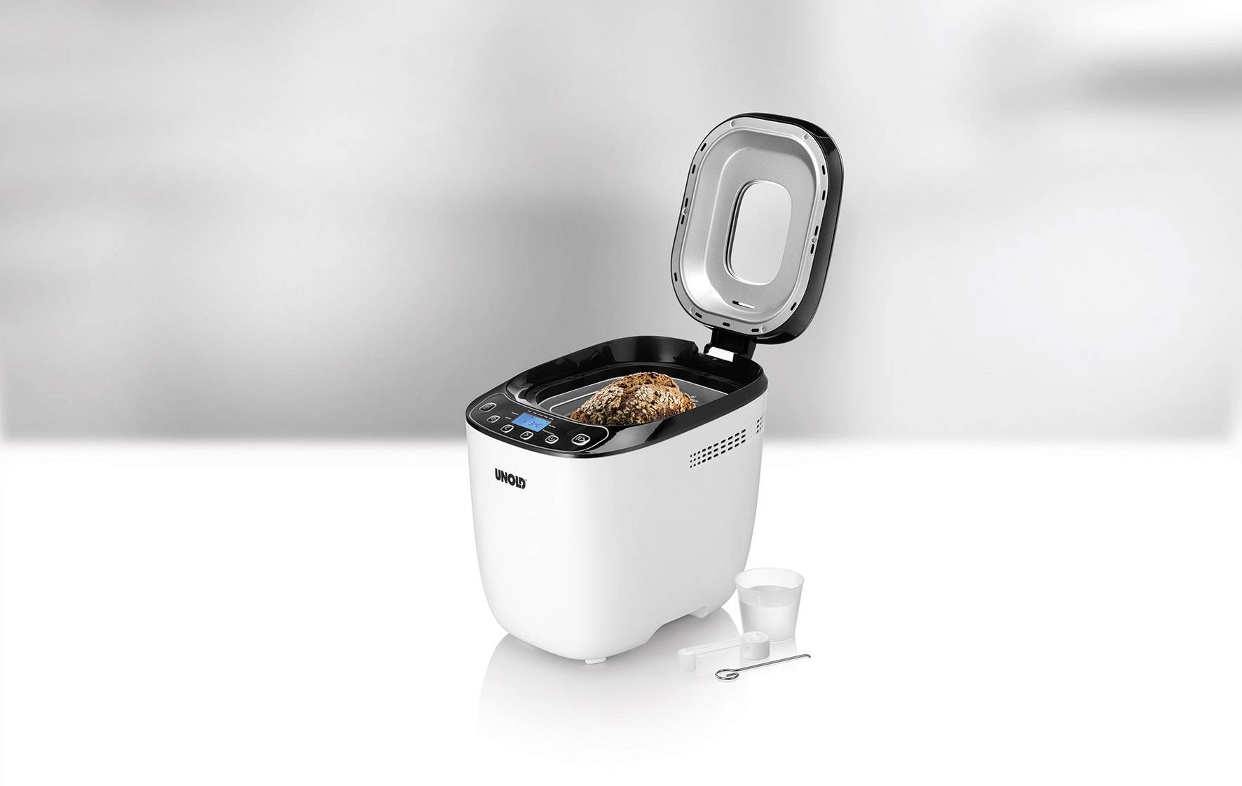 'Broodbakmachine met open deksel, toont een vers gebakken brood aan de binnenkant. Bijgevoegd een maatbeker en een kleine lepel.'