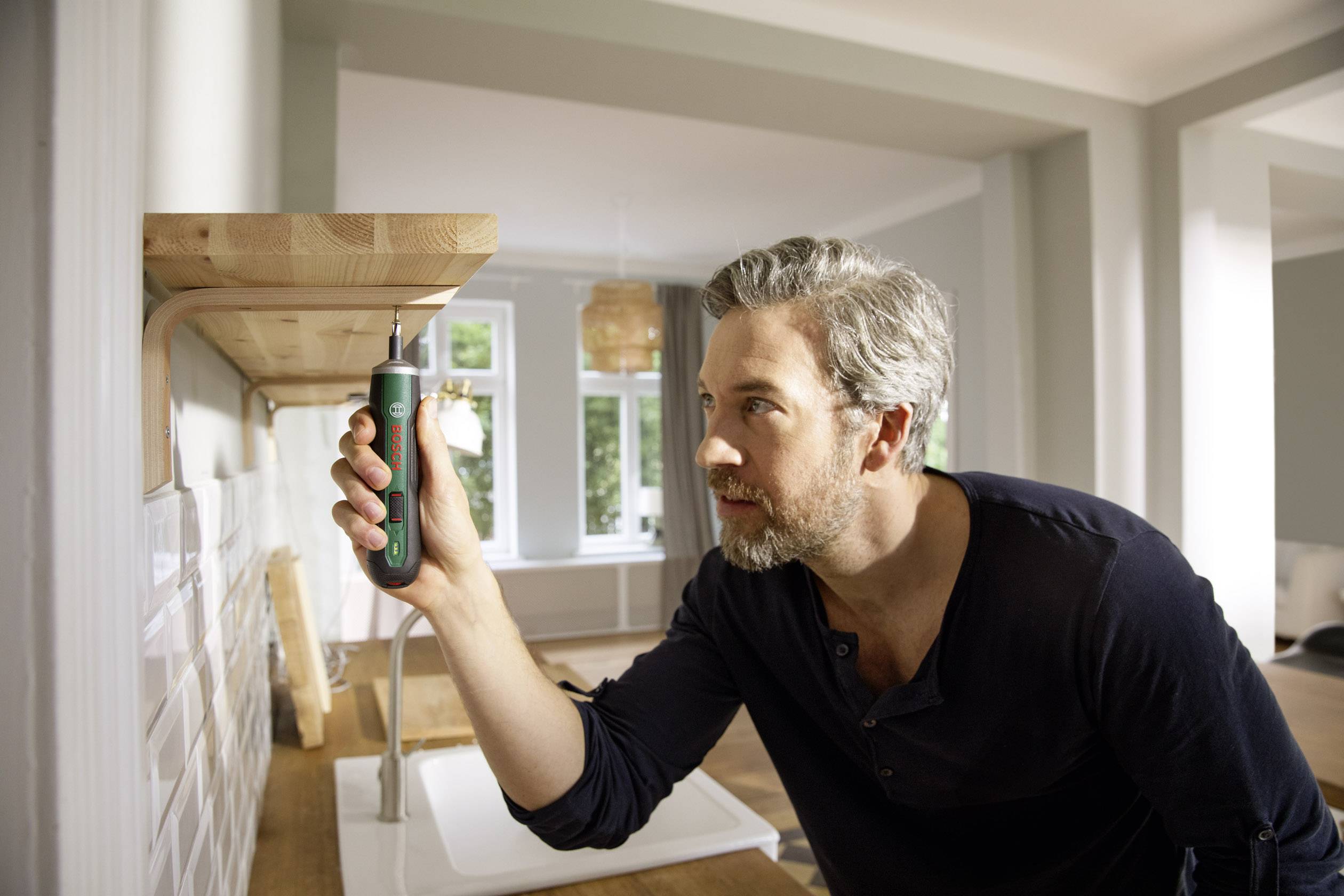 Een man monteert een houten rek aan de muur met een elektrische schroevendraaier. Op de achtergrond is een lichte woonkamer zichtbaar.