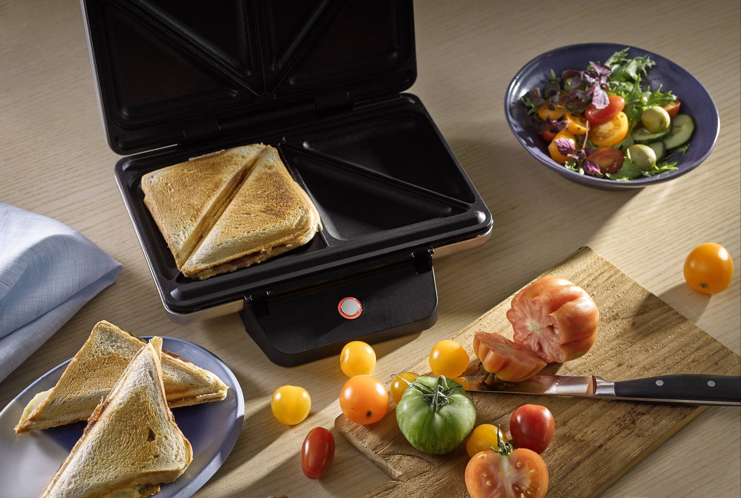 Een tostapparaat roostert een broodje. Ernaast ligt een houten plank met tomaten en een bord sla. Maaltijdvoorbereidingsscène.