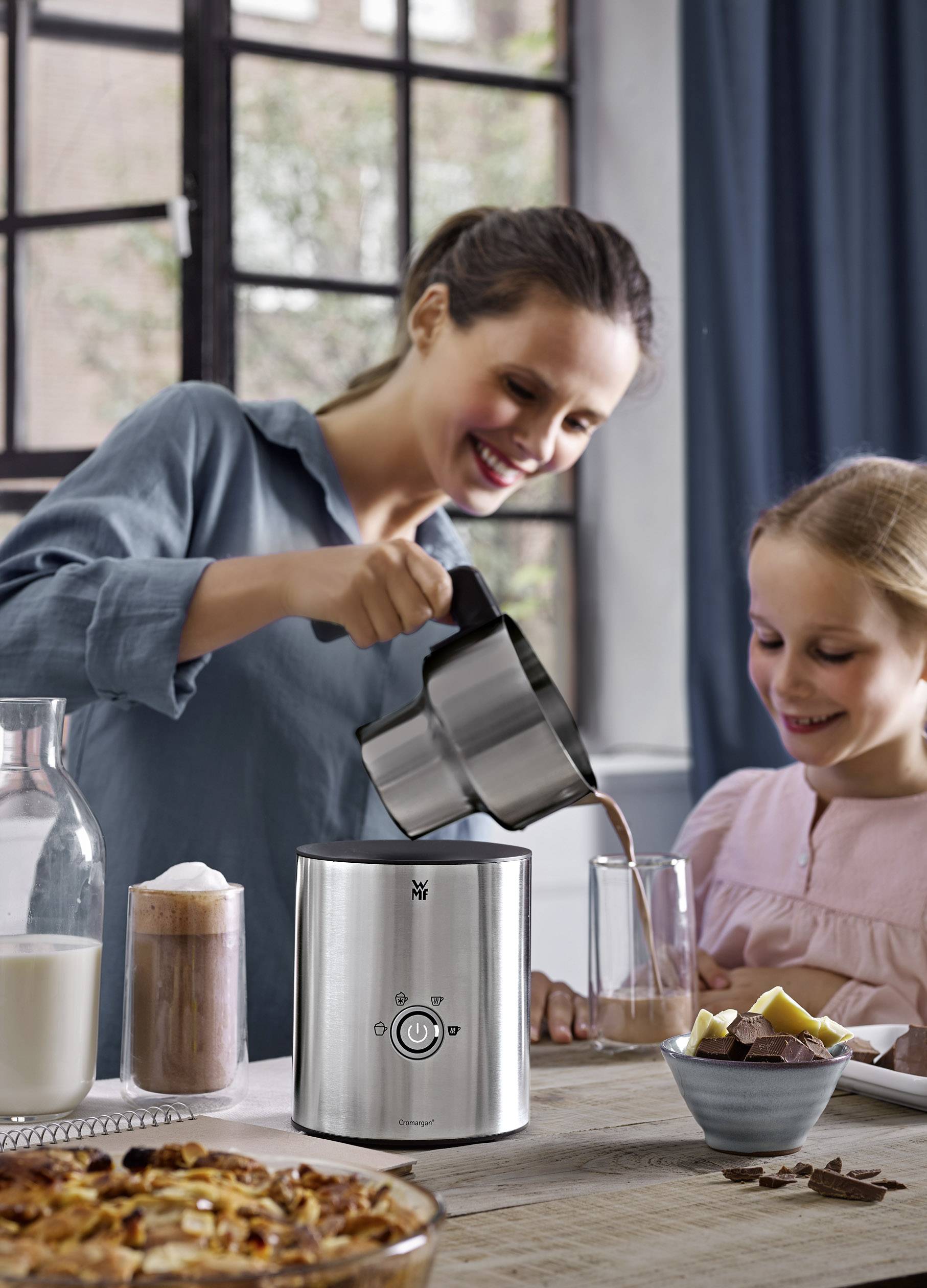 Een vrouw schenkt warme chocolademelk uit een melkopschuimer in een glas, terwijl een meisje toekijkt. Op de achtergrond een uitzicht op het raam.