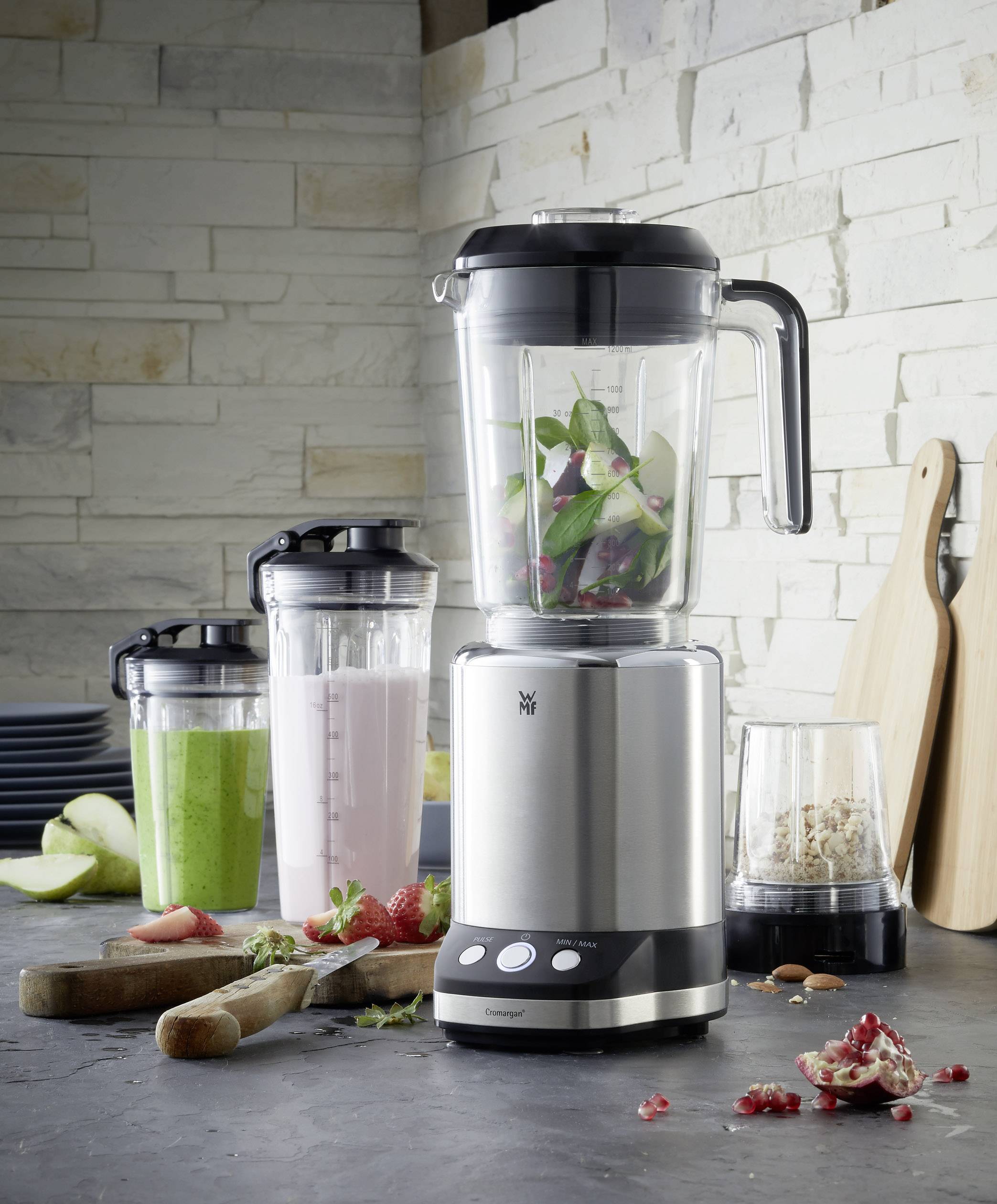 Een zilveren blender op een aanrecht, gevuld met groen groente. Ernaast staan smoothies, een snijplank en ingrediënten.