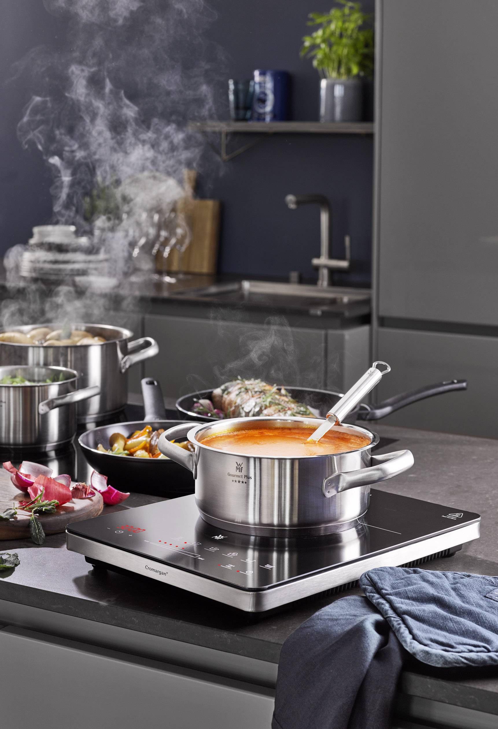 Een pan met kokende soep op een fornuis in een moderne keuken. Stoom stijgt op, groenten en kruiden liggen ernaast.