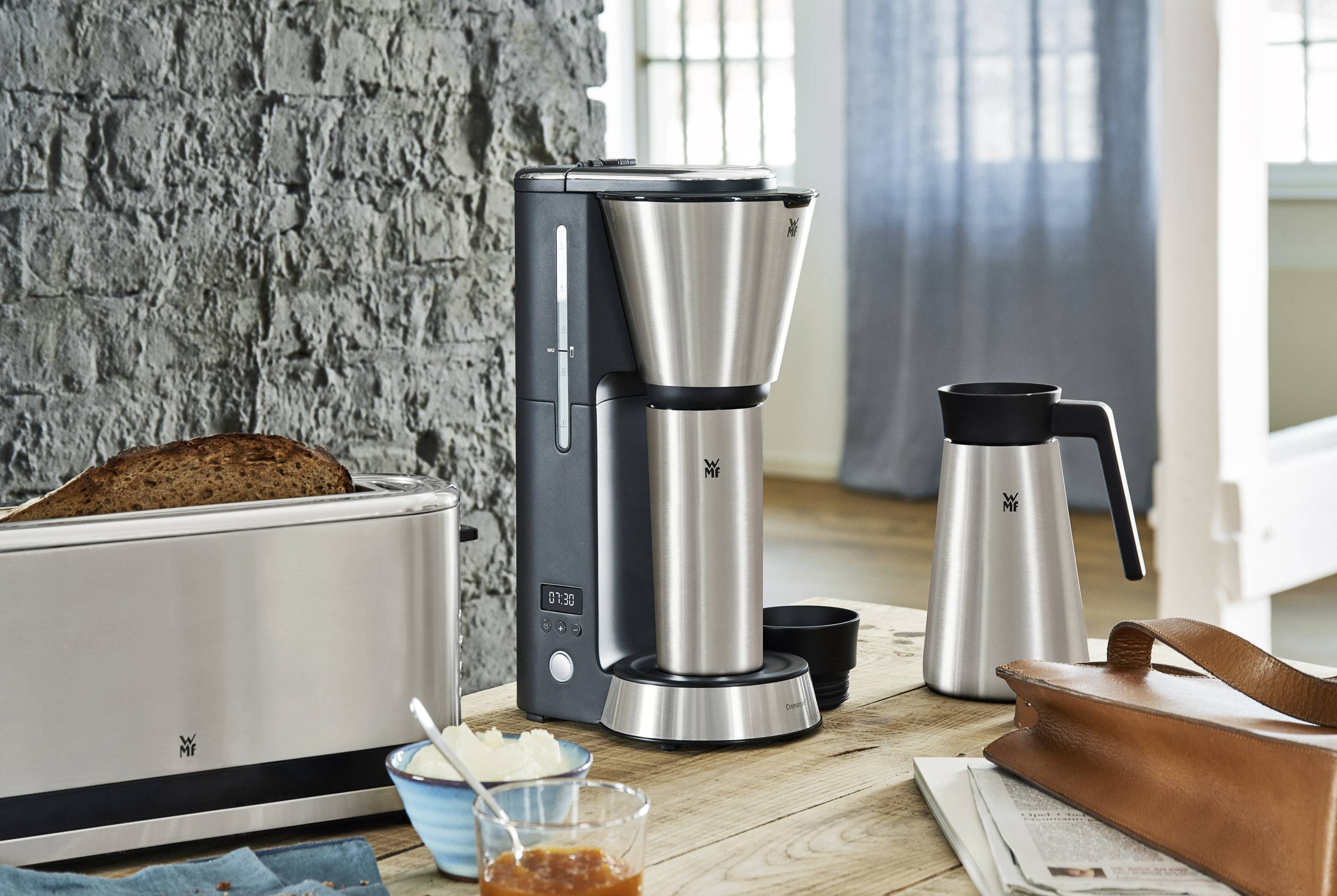 Een koffiemachine van roestvrij staal staat op een houten tafel naast een broodrooster, een thermoskan en een bruine leren tas.