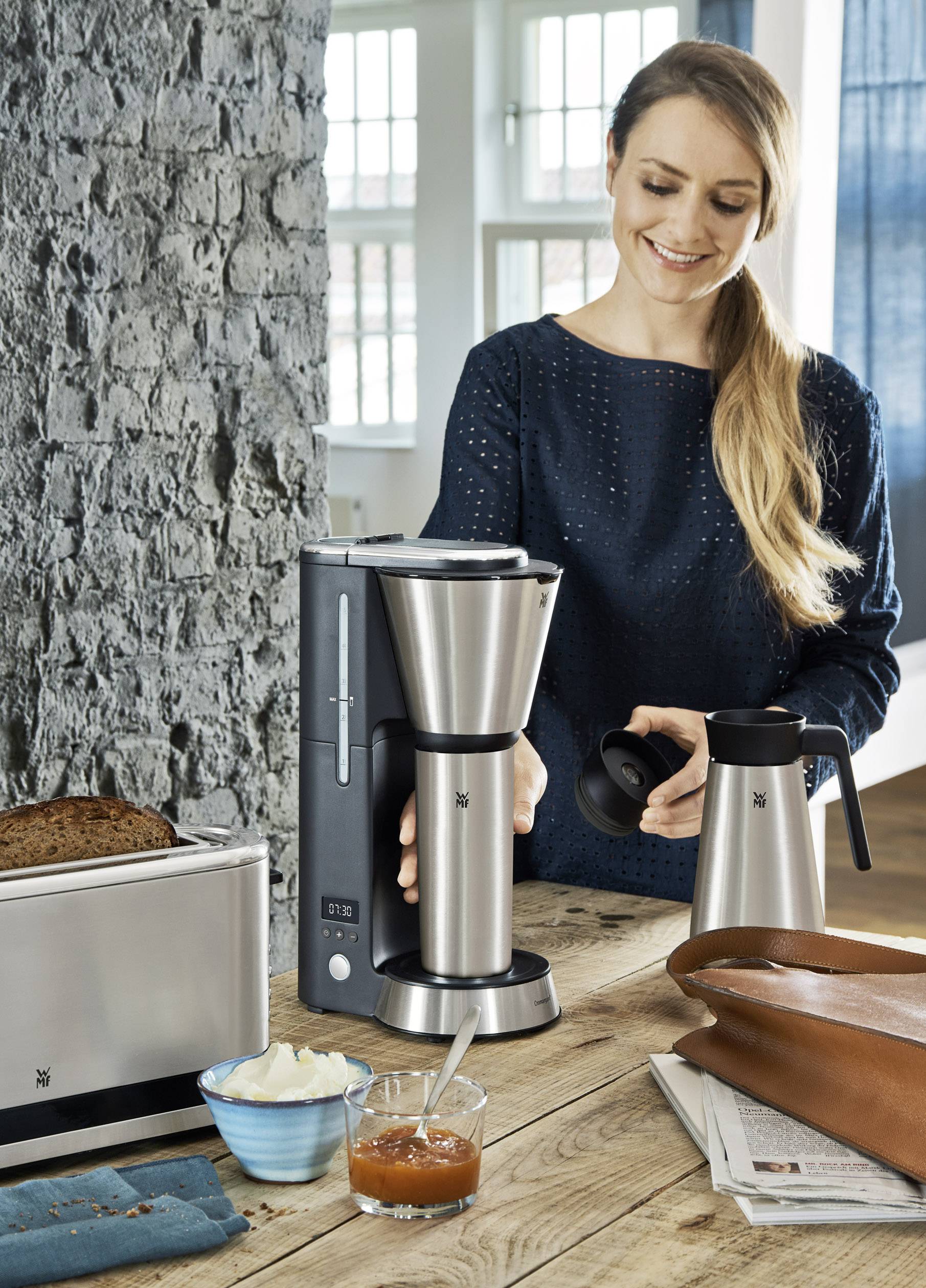 Een vrouw staat in een keuken en bereidt koffie met een zilveren koffiemachine. Op tafel liggen brood en jam.