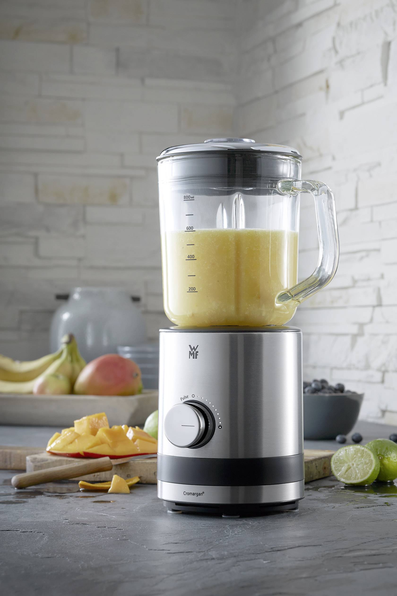 Een zilveren staande blender met glazen kan bevat een oranje smoothie. Op de achtergrond liggen vruchten op een snijplank.