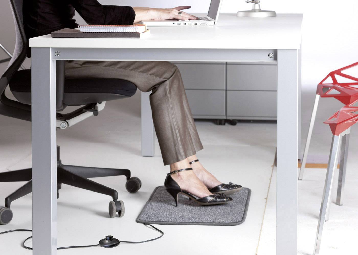 Een persoon zit aan een bureau en werkt op een laptop. Zijn/haar voeten rusten op een grijze voetenmat onder de tafel.