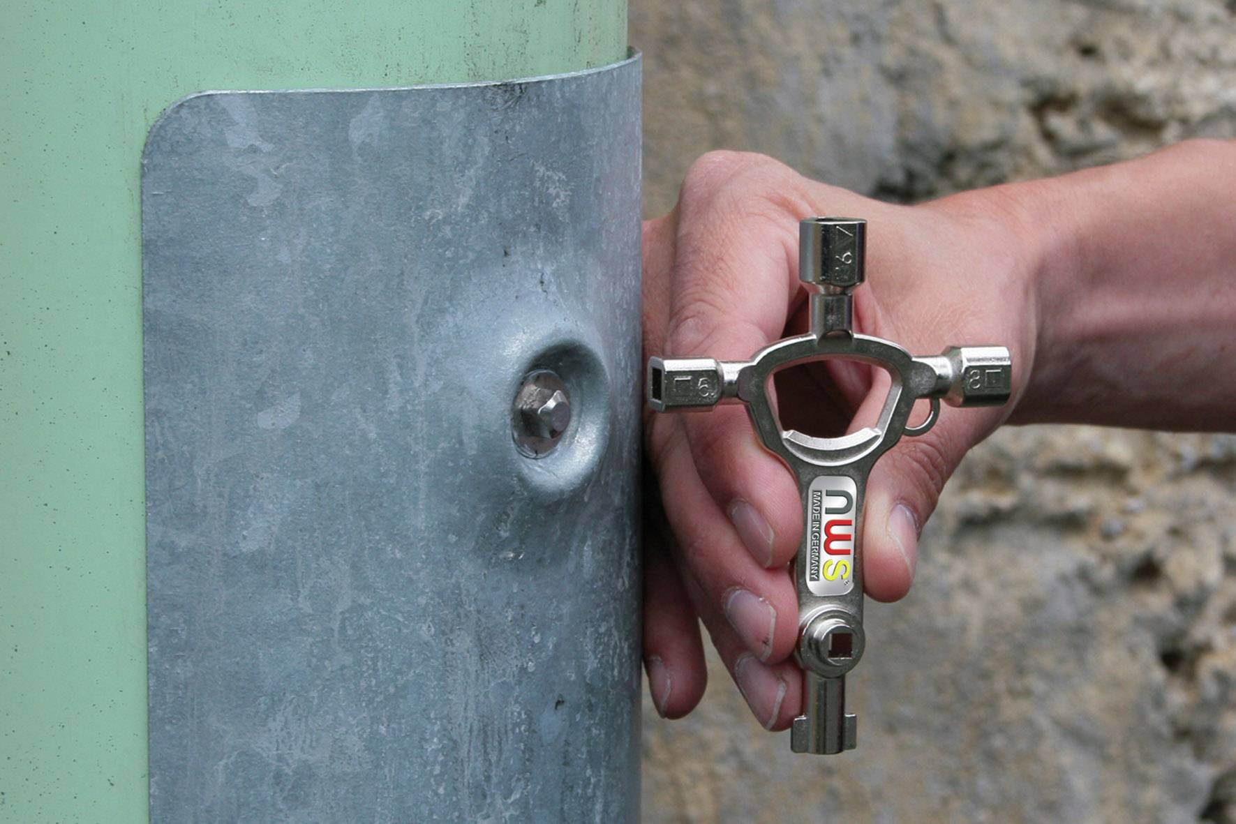 Een hand houdt een vierkante sleutel vast om een metalen behuizing aan een groene zuil te openen. De behuizing heeft een schroefopening.