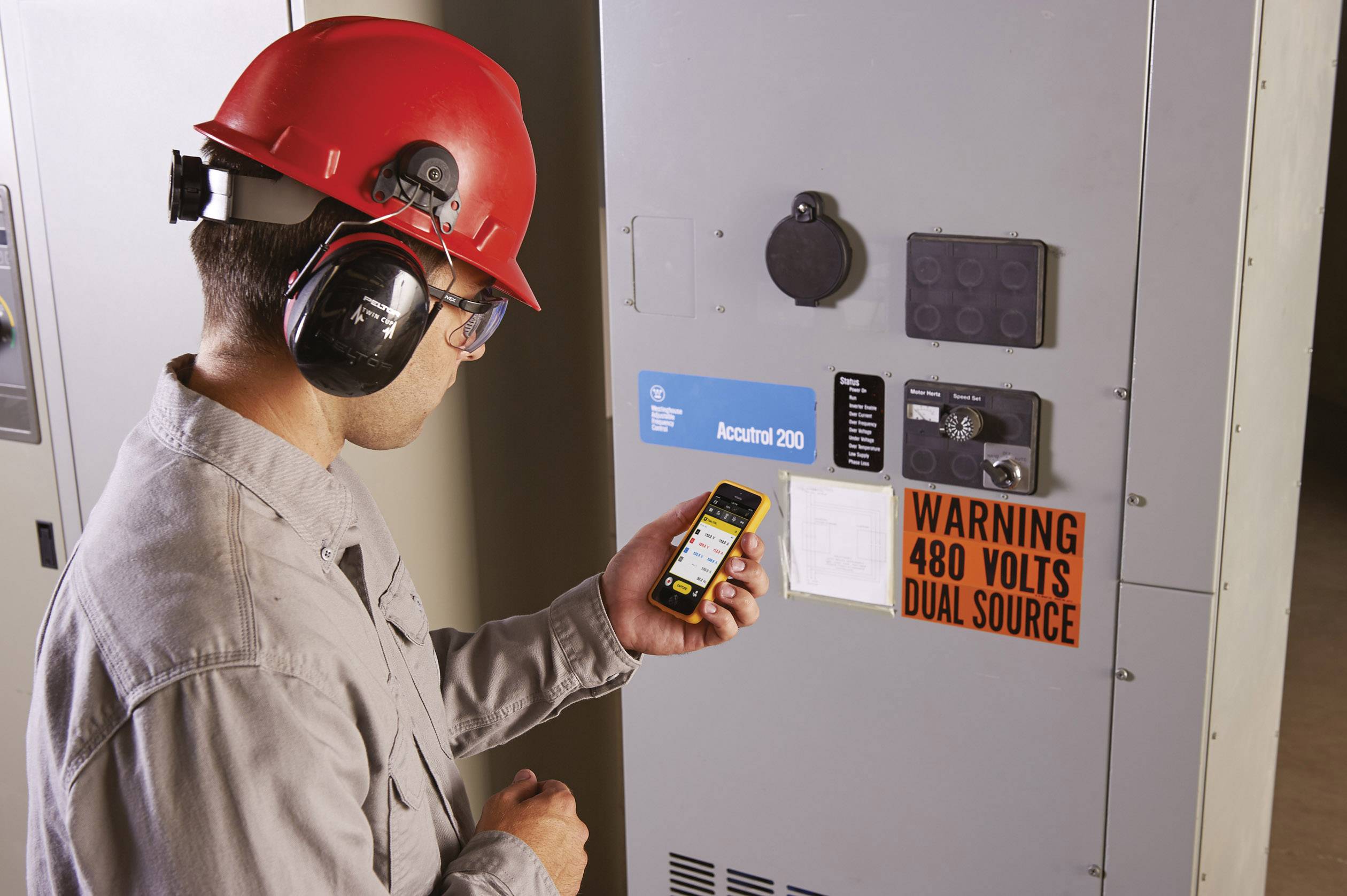 Een arbeider met een rode veiligheidshelm en gehoorbescherming staat voor een elektriciteitskast en leest op een smartphone. Naast hem hangt een waarschuwingsbord.