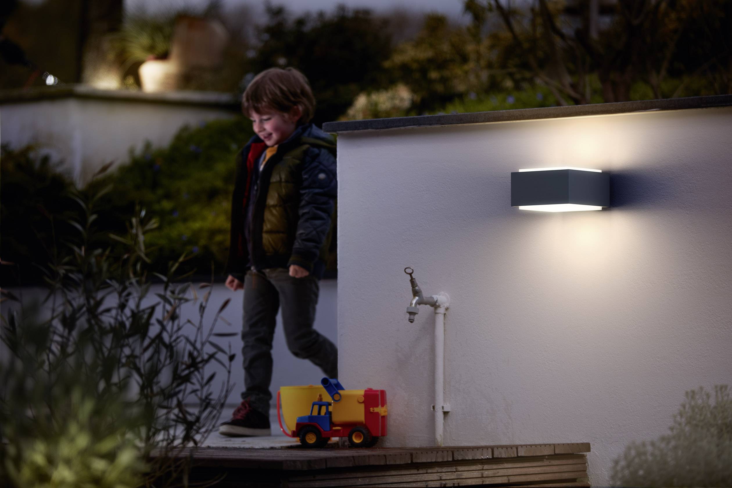 Een kind speelt buiten naast een wandlamp en een speelgoed-vrachtwagen. De omgeving is zacht verlicht en heeft een avondlijke sfeer.
