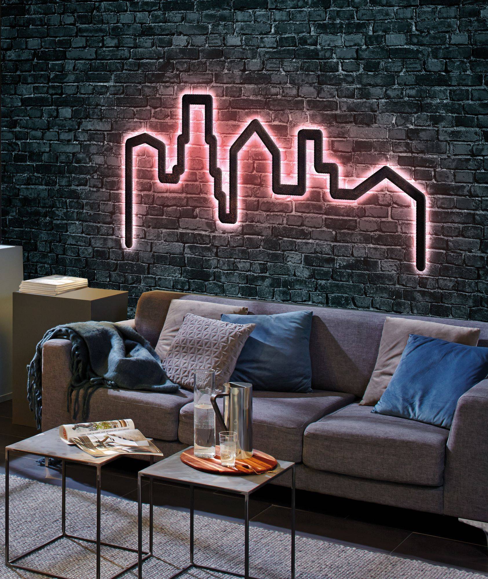Woonkamer met een grijze bank, blauwe sierkussens en een salontafel. Aan de achterwand een roze verlicht wandkunstwerk met een stadssilhouet.