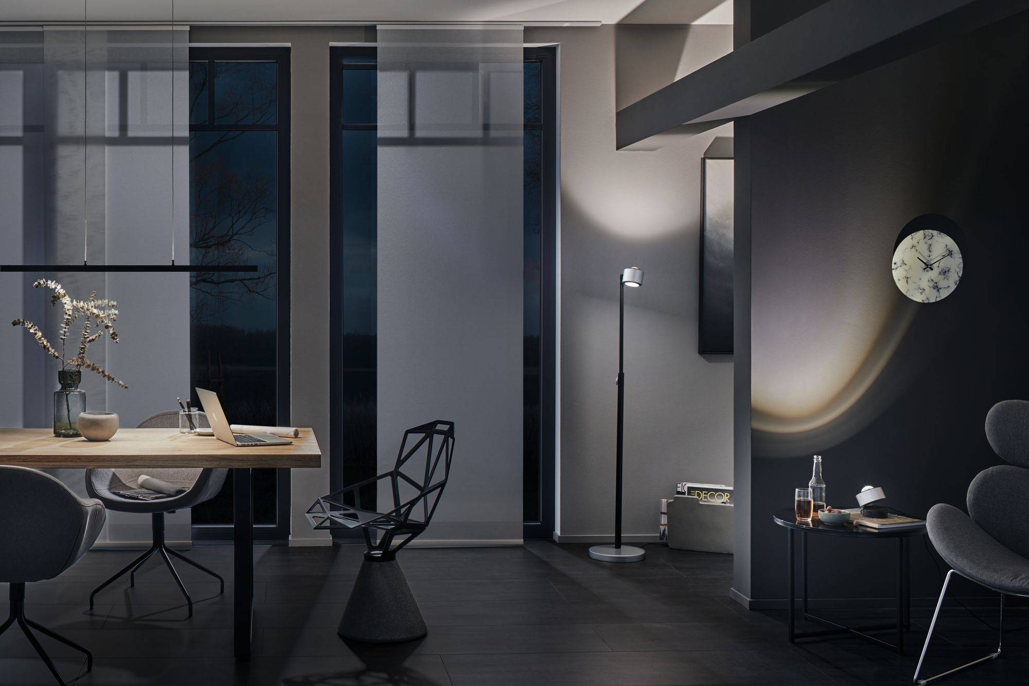 Moderne woonruimte 's avonds met indirecte verlichting, grote tafel, laptop en decoratieve stoel. Lichte muren, klok aan de muur.