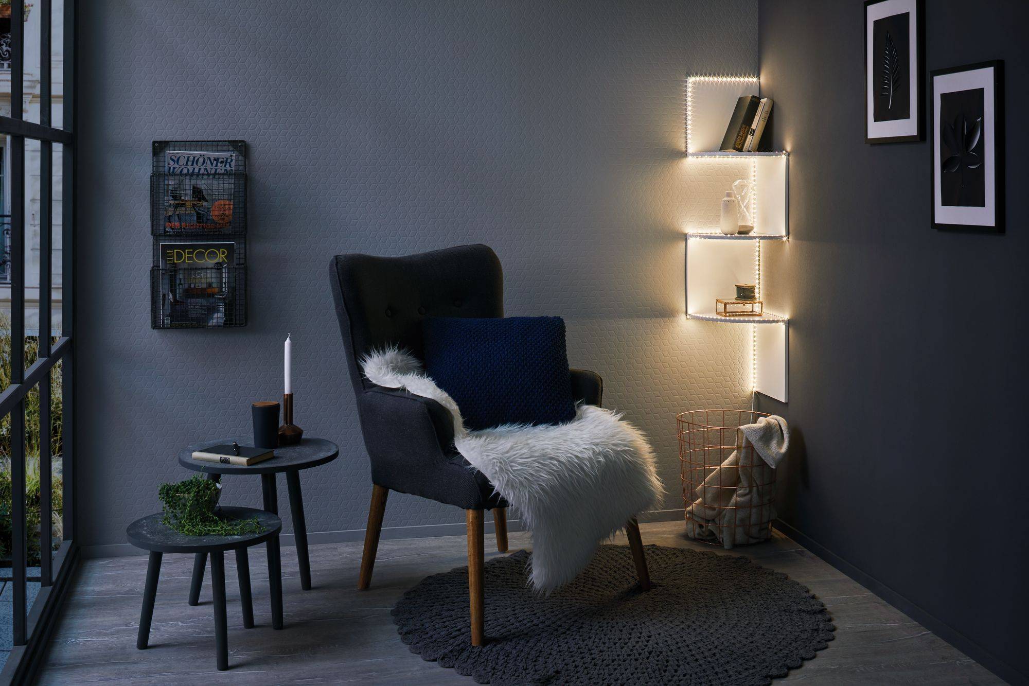 Gezellige leeshoek met een grijze fauteuil, blauwe kussens en een wit vacht. Verlicht rek aan de muur. Moderne decoratie.