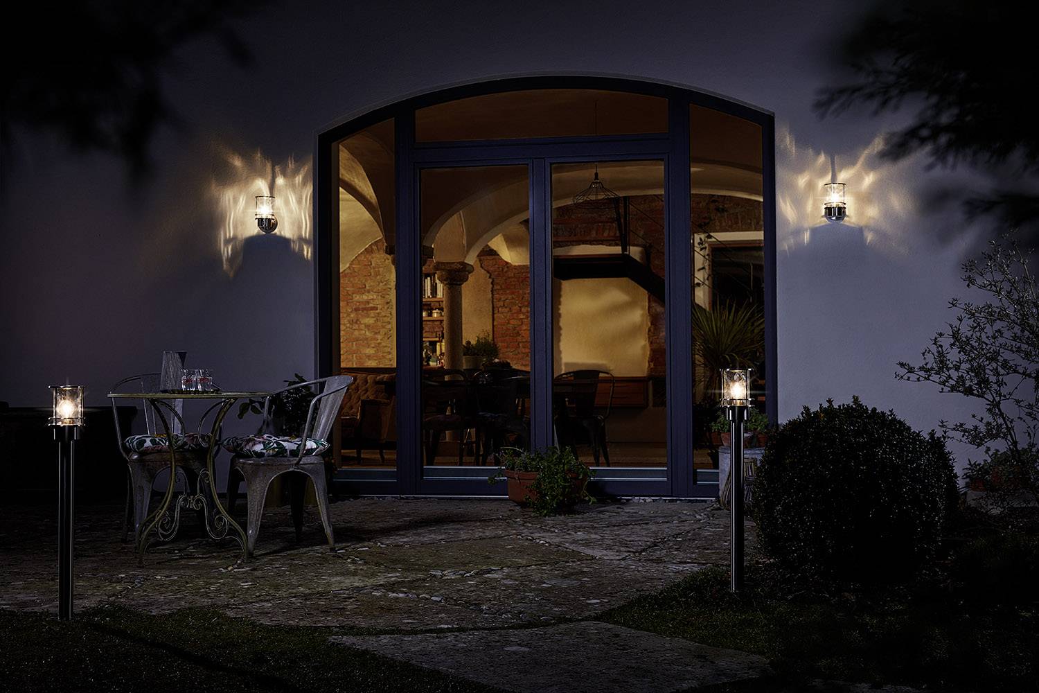 Tuin bij het huis 's nachts met verlicht raam. Links staat een tafel met stoelen, en op de achtergrond is een warm verlichte woonkamer zichtbaar.