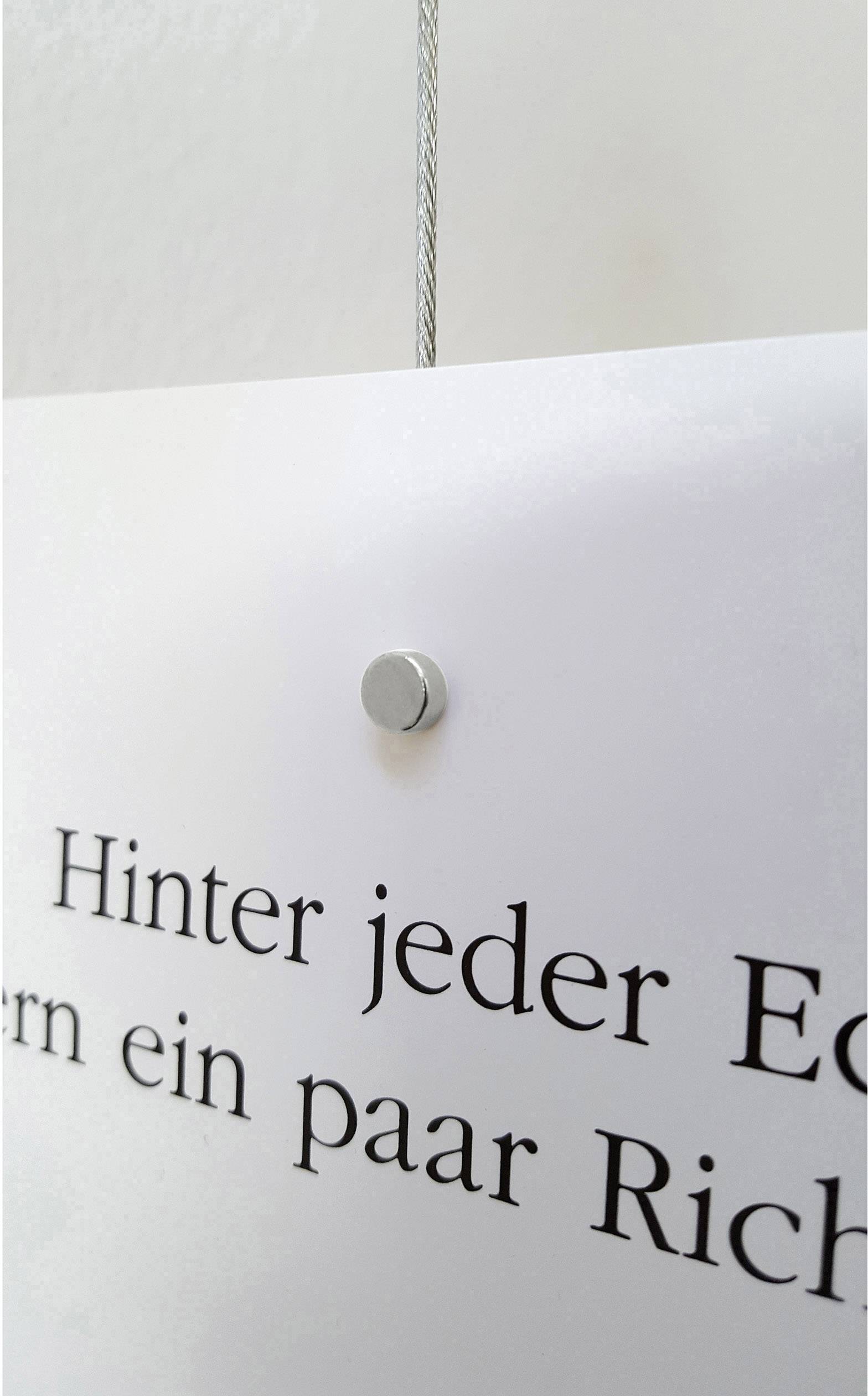 Een wit bord hangt aan een staaldraad. Het toont gedeeltelijk de tekst 'Achter elke hoek een paar Rich' in zwarte letters.