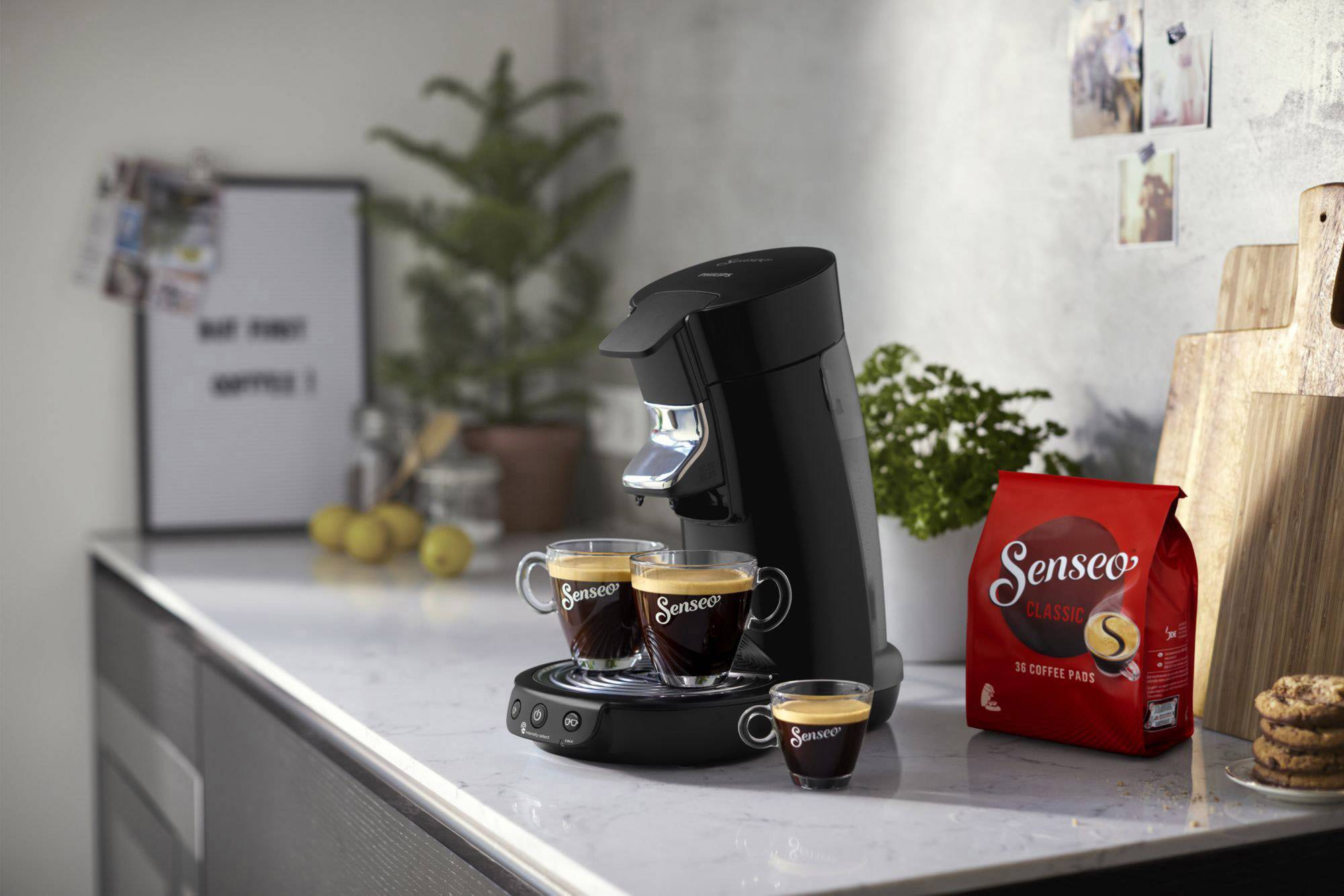 Een zwarte koffiemachine zet twee kopjes koffie klaar op een aanrecht. Ernaast staat een pak koffiepads.