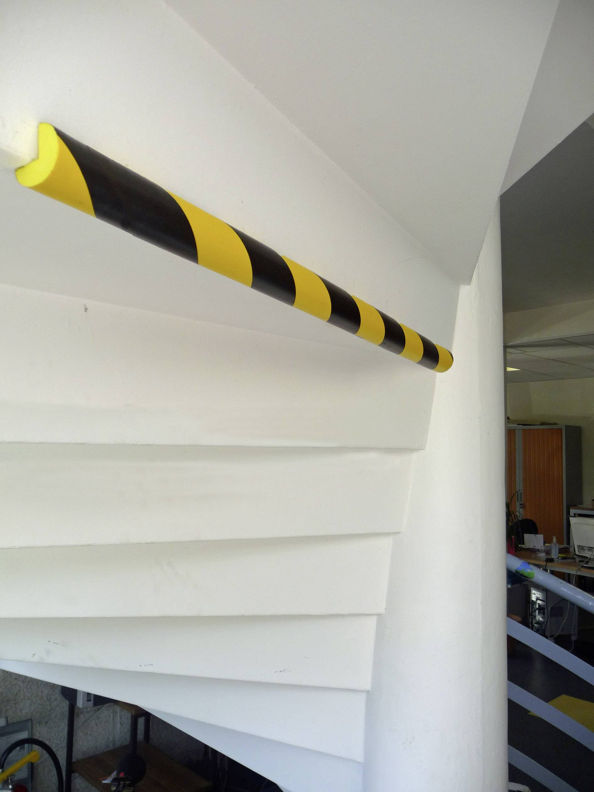 Geel-zwarte waarschuwingsmarkering op de traprand in een binnenruimte, bedoeld om botsingen te voorkomen. Kantooromgeving zichtbaar op de achtergrond.