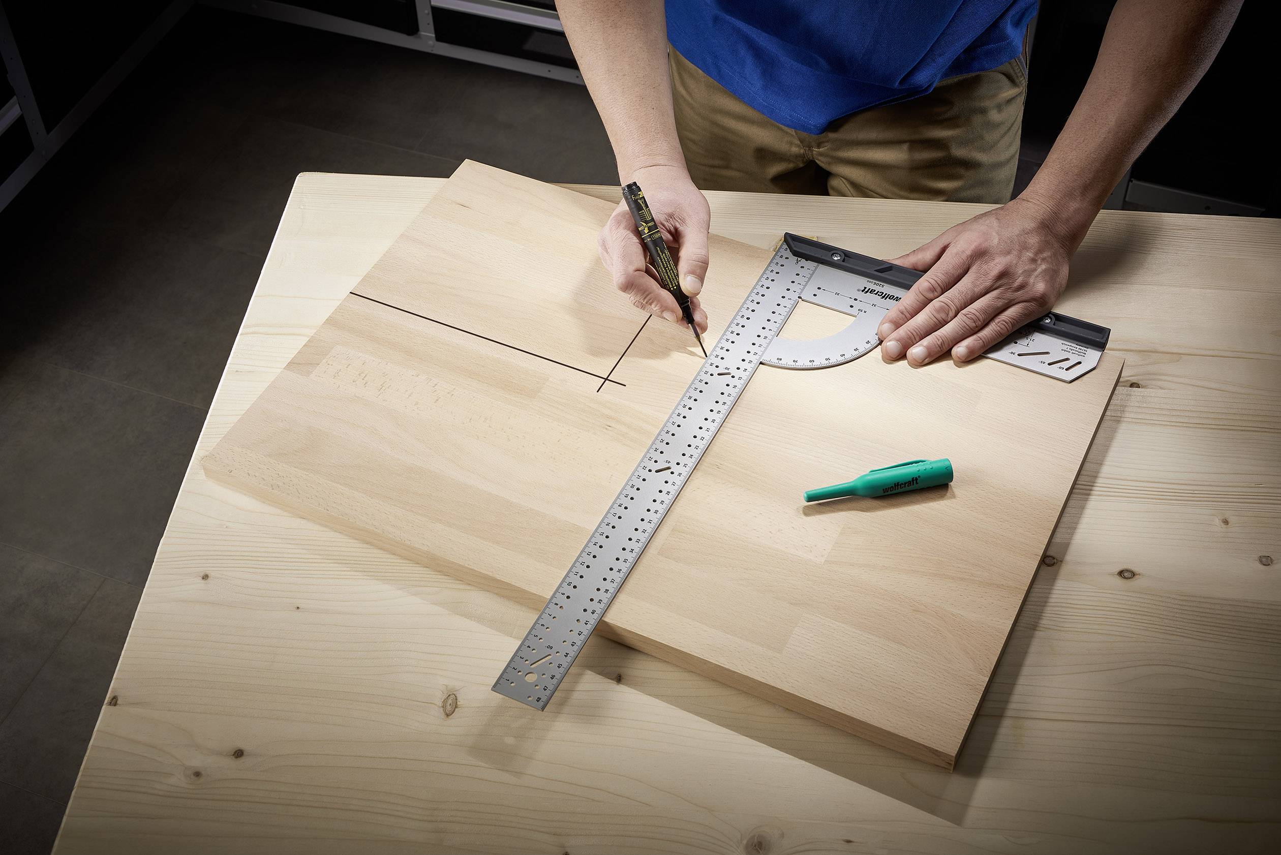 Een persoon meet een houten plank op een houten tafel met een geodriehoek en potlood. Een groene pen ligt ernaast.