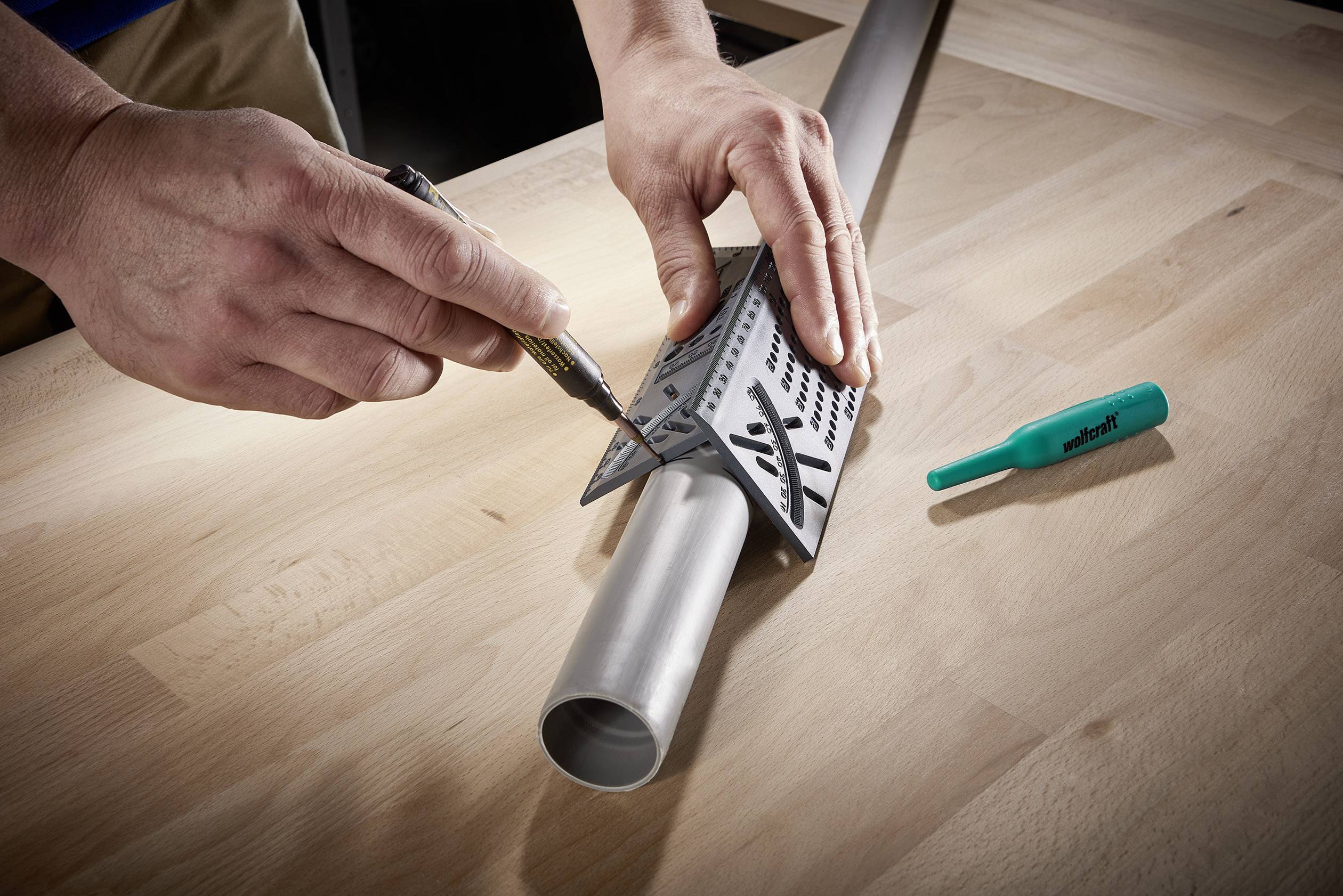 Een persoon meet met een meetlat en pen afmetingen op een metalen buis die op een houten tafel ligt. Een groene pen ligt ernaast.