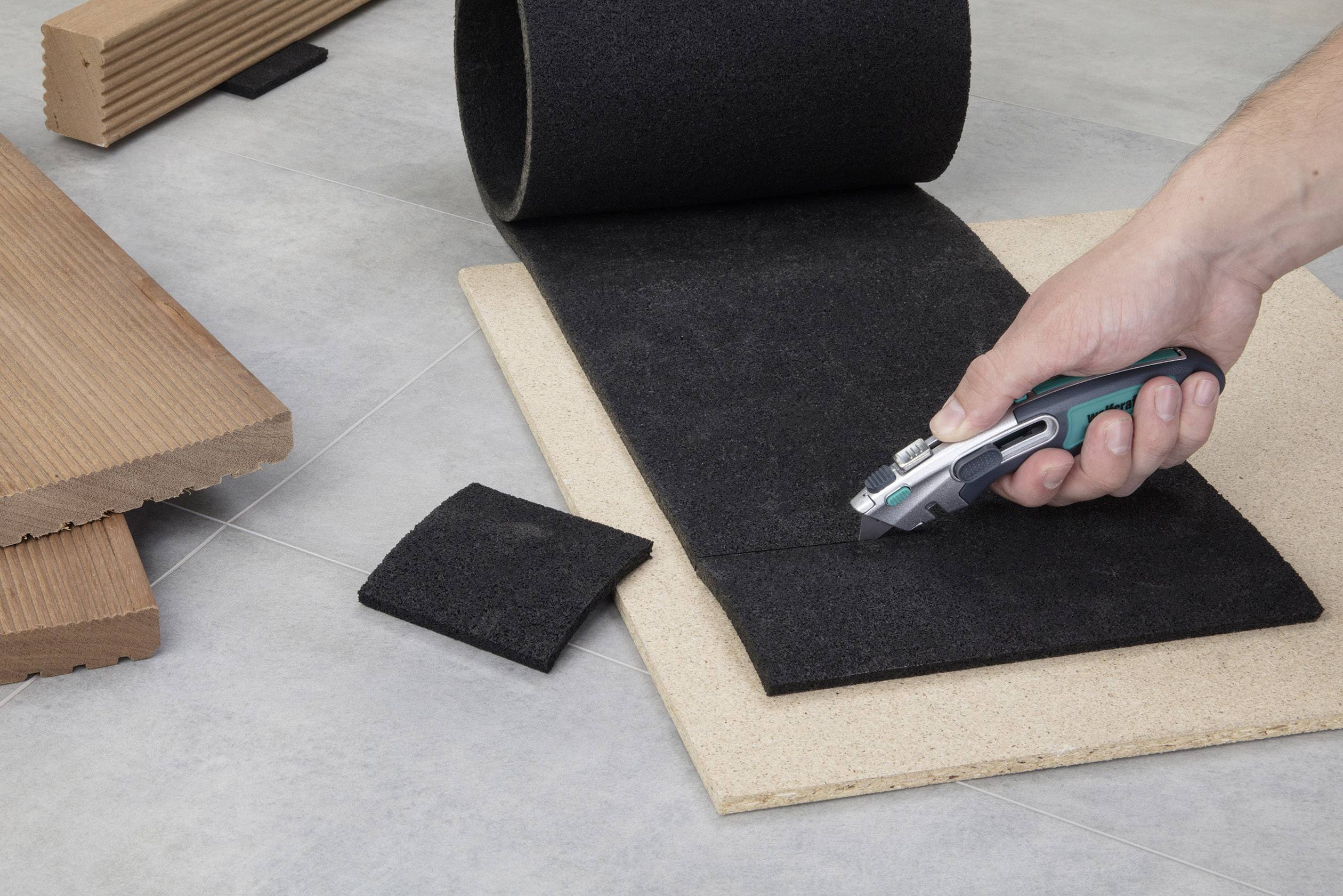 Een hand snijdt met een tapijtmes een stuk zwarte vloerbedekking op een ondergrond, omringd door hout- en tapijtrestanten.