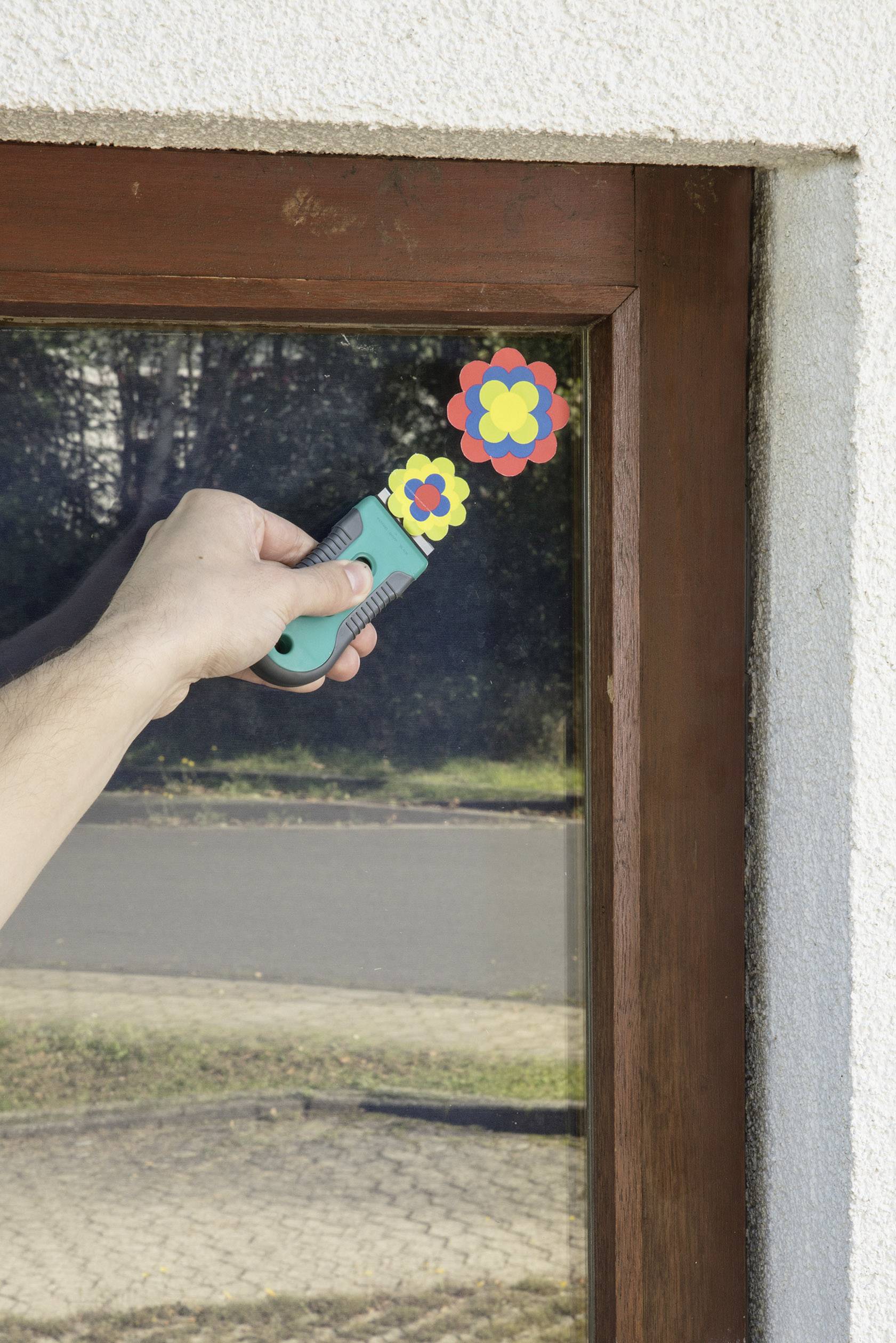 Een hand houdt een raamreiniger vast, terwijl kleurrijke bloemen stickers op een raam zijn aangebracht.
