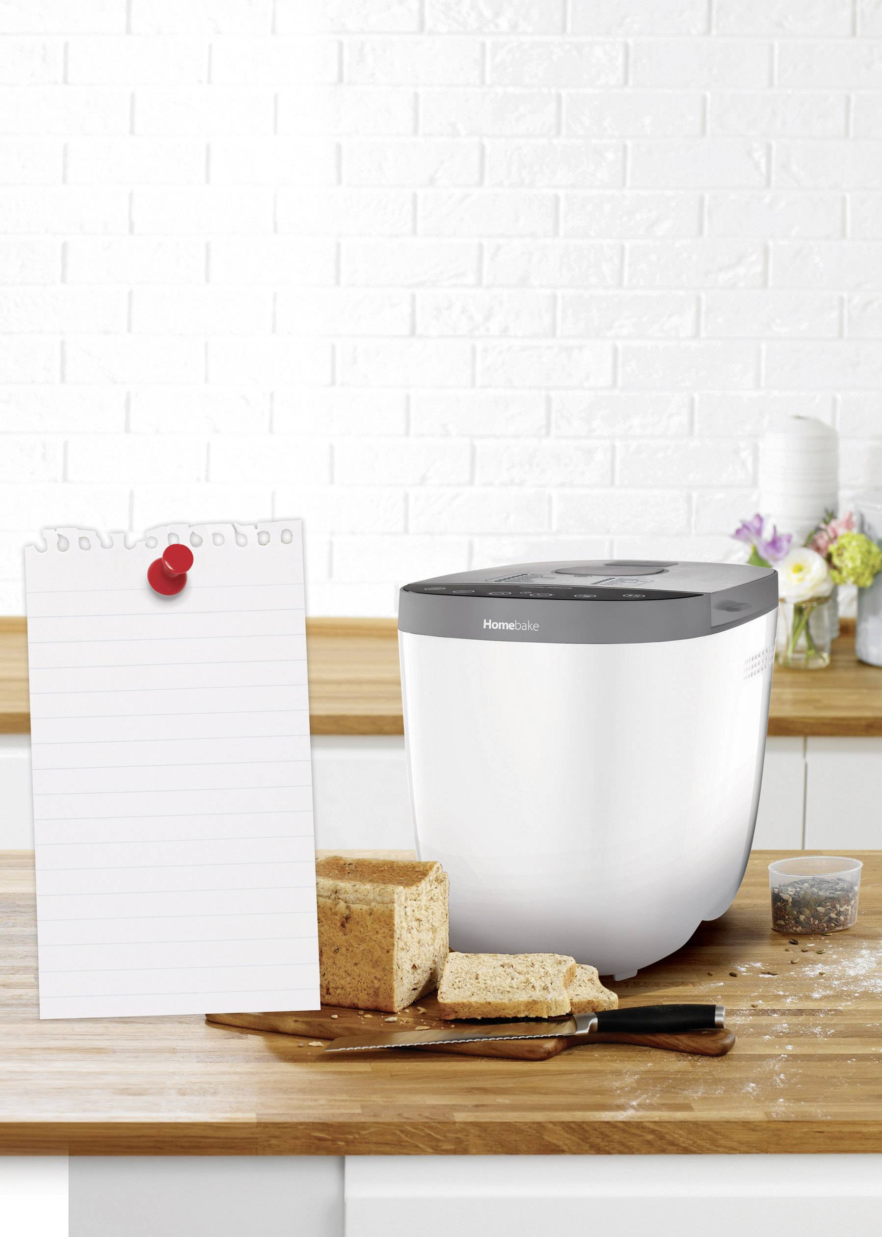 Een broodbakmachine op een aanrecht met een gebakken brood ernaast. Op de achtergrond een witte bakstenen muur en bloemen.