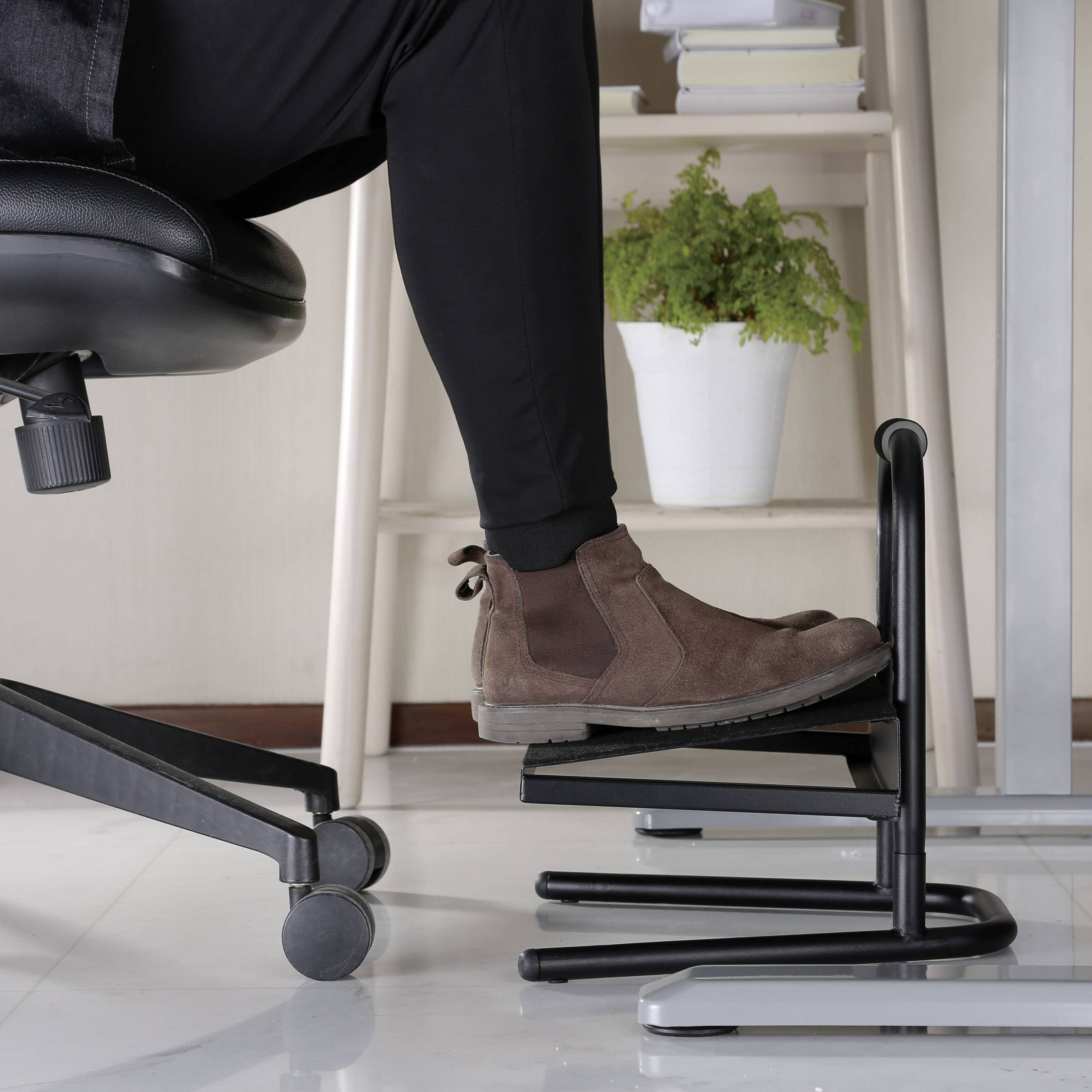 Een been in een bruine schoen op een ergonomische voetensteun onder een bureaustoel naast een kleine tafel met planten.