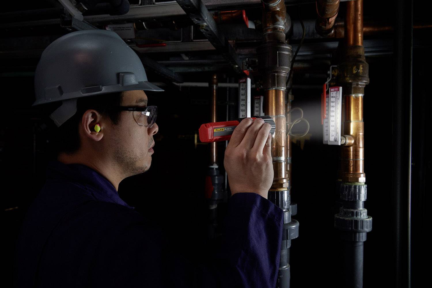 Een persoon met een veiligheidshelm en beschermende bril inspecteert buizen in een industriële omgeving met een rood apparaat voor het detecteren van gaslekken.