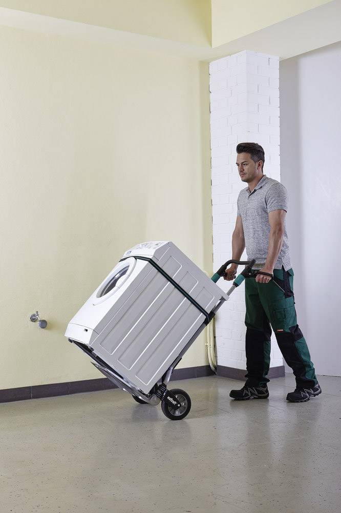 Een man in werkkleding vervoert een wasmachine met een steekwagen. Hij beweegt zich in een ruimte met lichte muren.
