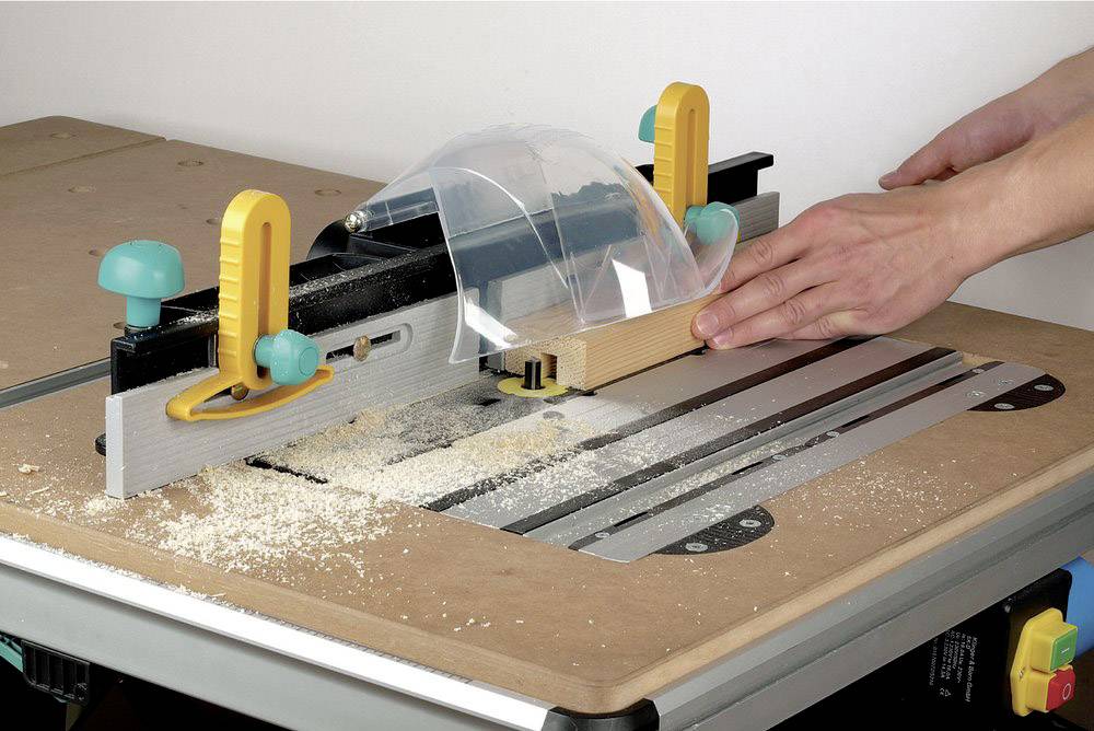 Een persoon geleidt een houten plank door een tafelfreesmachine met een beschermkap. Houtspaanders zijn rond de machine verspreid.