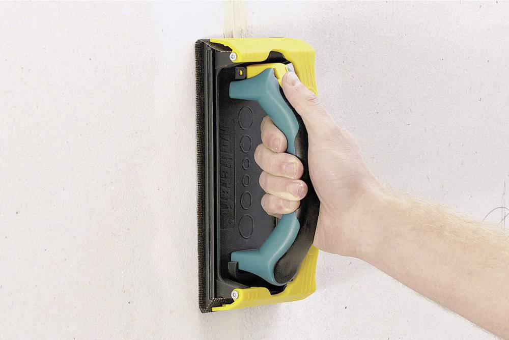 Een hand houdt een gele schuurblok met een zwart schuurvlak tegen een witte muur. Schuurbeweging wordt gesimuleerd.