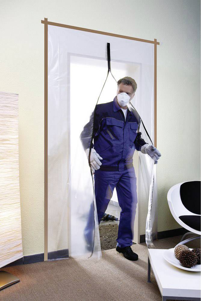 Een man in beschermende kleding en een masker betreedt een ruimte door een transparante plastic barrière.