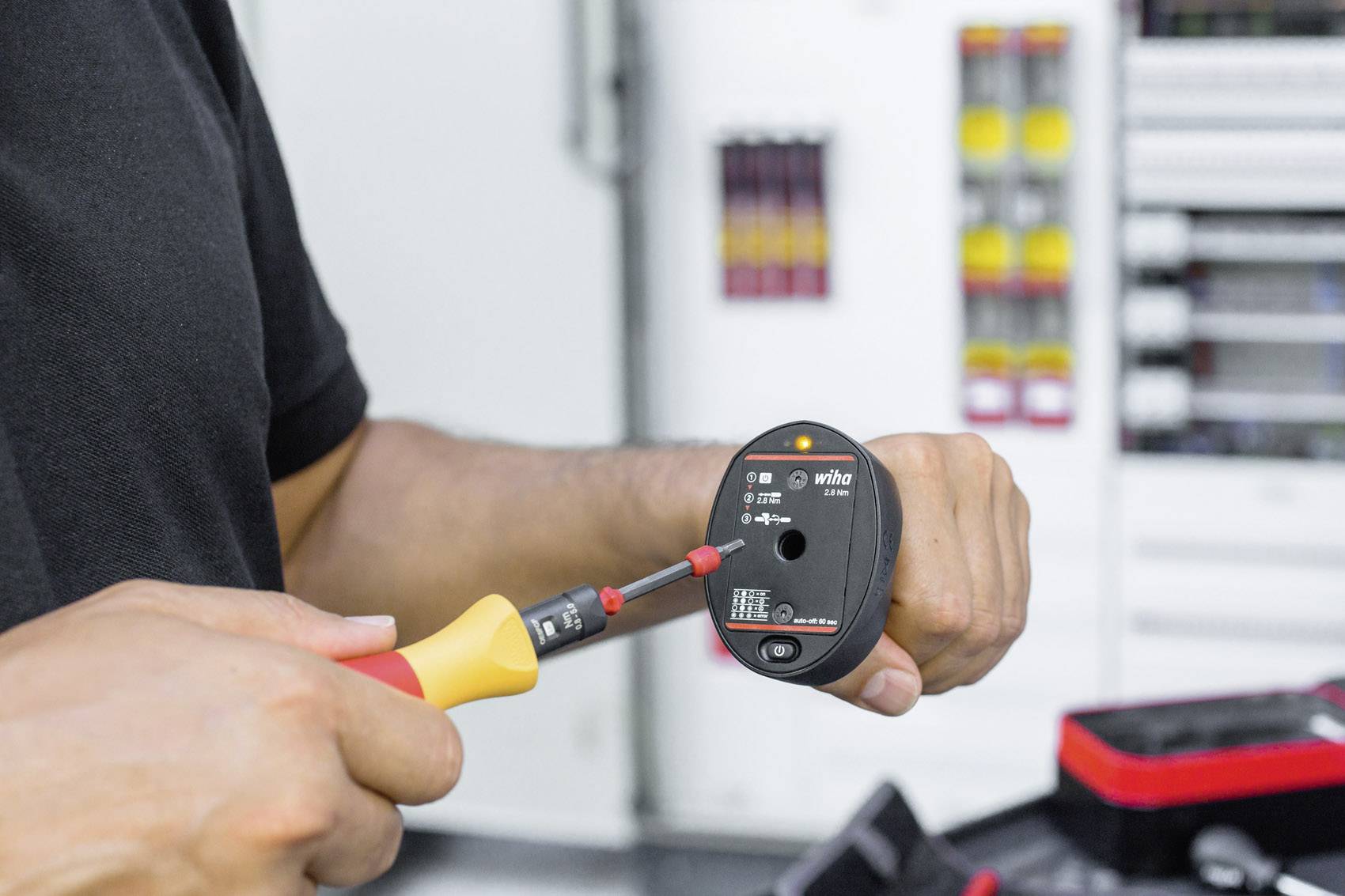 Een persoon houdt een elektrisch meettoestel vast en test een spanning. Op de achtergrond zijn onscherpe schakelkasten te zien.