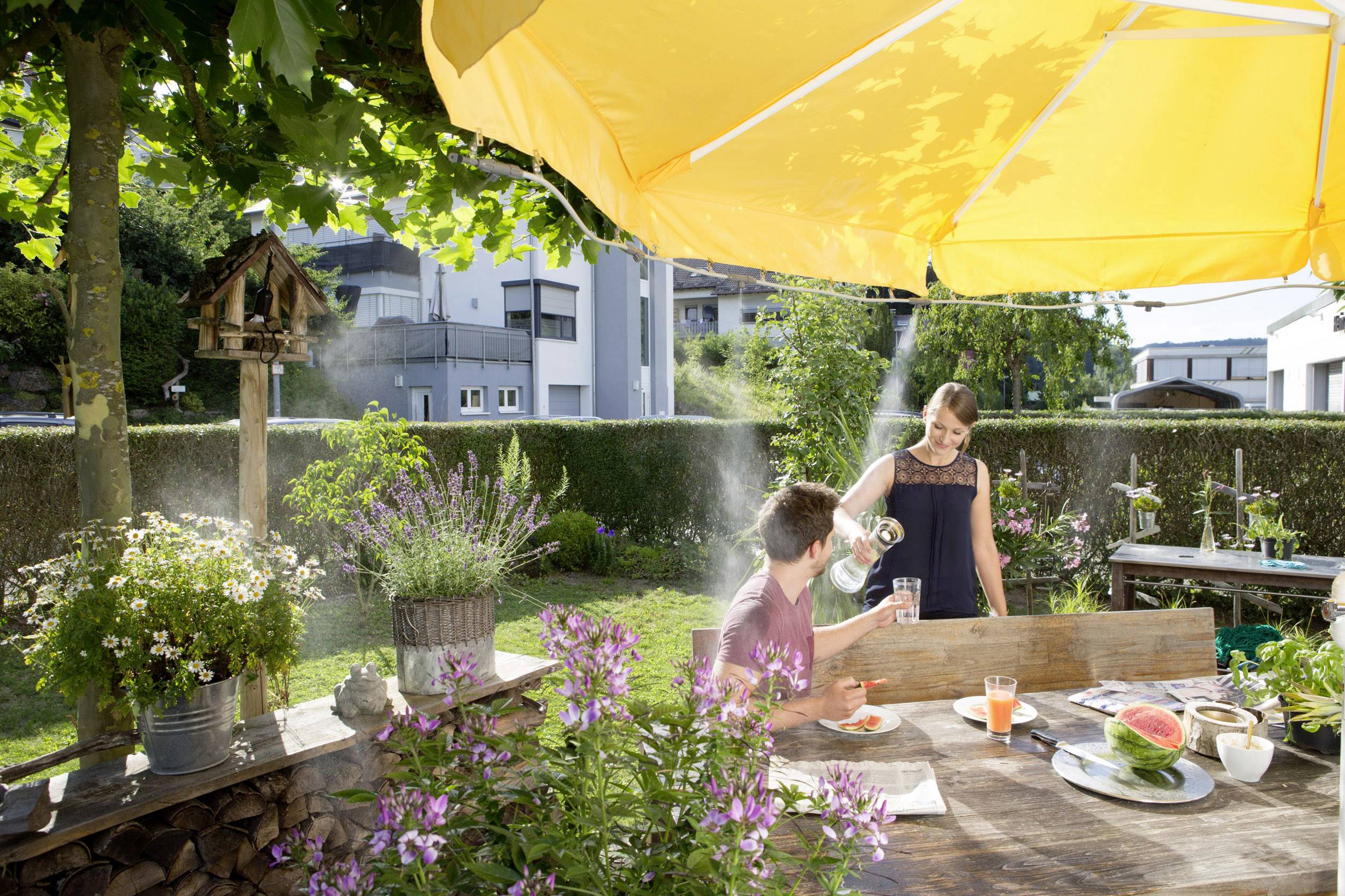 Een man en een vrouw zitten in de zonnige tuin aan een houten tafel. Hij krijgt water ingeschonken. Om hen heen bloeien planten.
