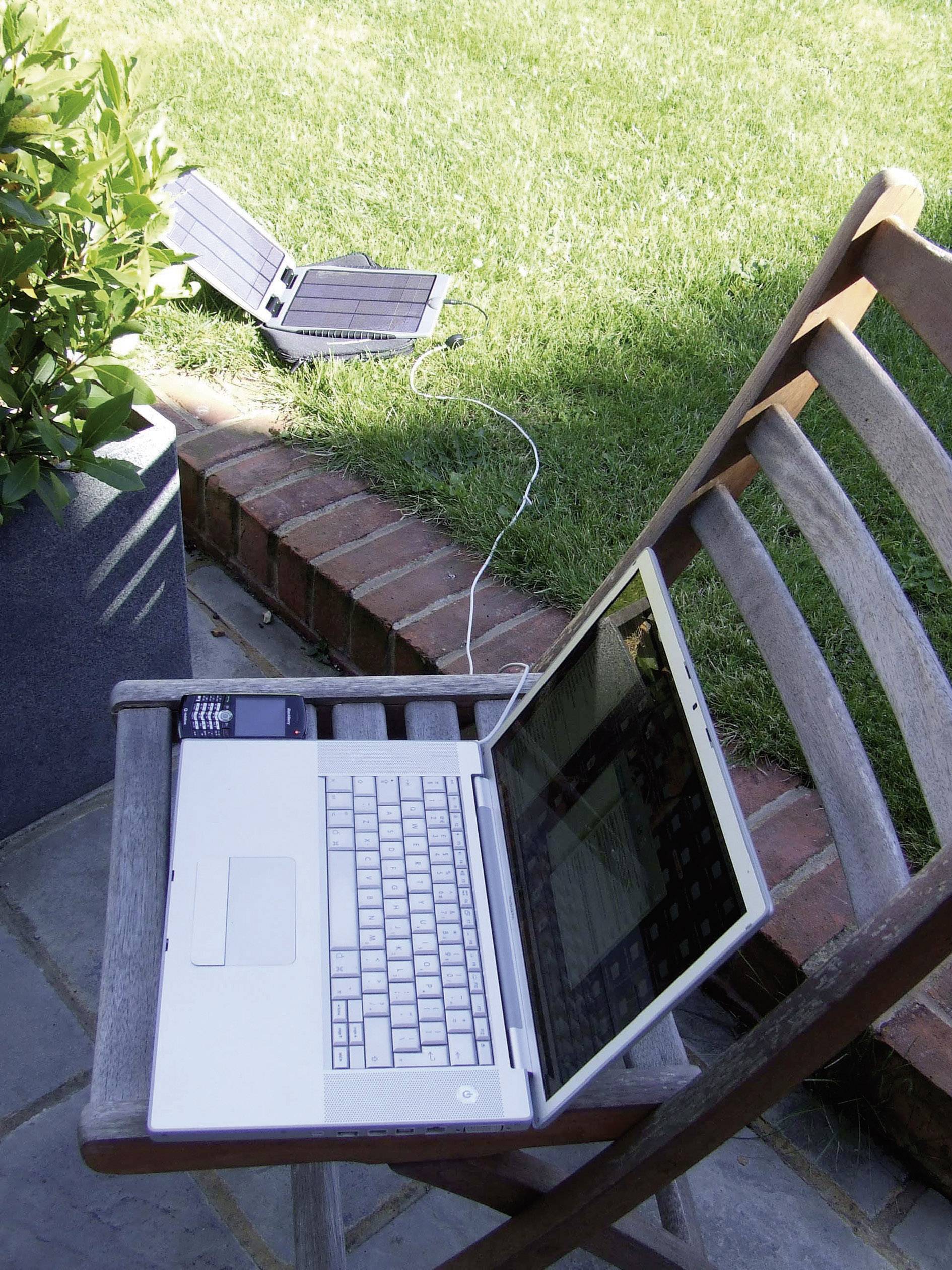 Een laptop op een houten stoel buiten, verbonden met een zonnepaneel op het gazon, demonstreert milieuvriendelijke energie.