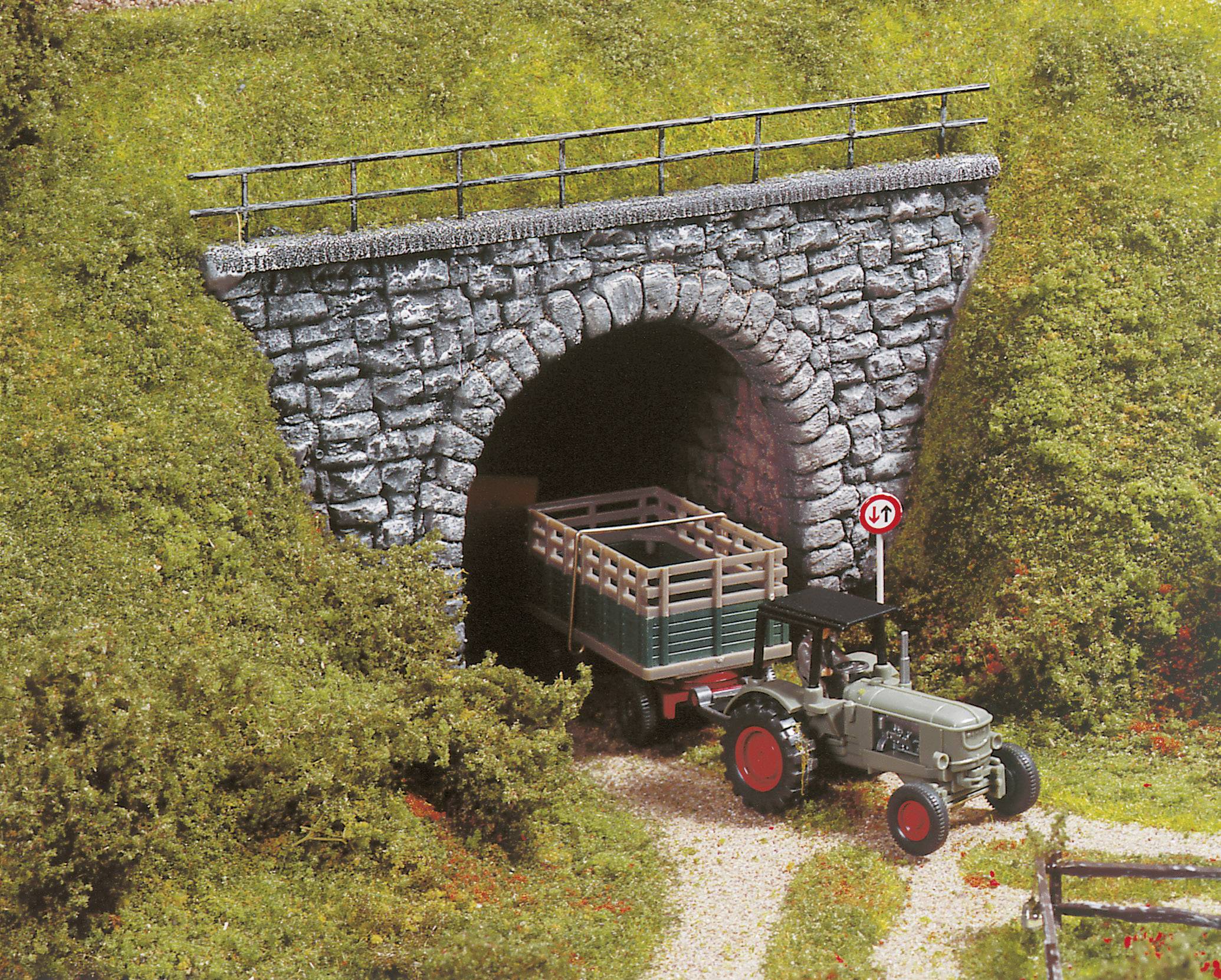 Een modeltraktor trekt een aanhanger uit een kleine steentunnel met een snelheidsbord '15'. De scène toont een landelijk terrein.