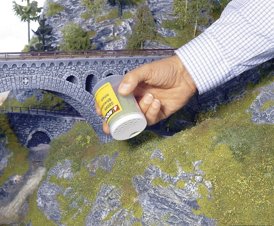 Een hand strooit grassimulatie uit een strooier over een modelspoorbaan-landschap met een brug en rotsen om de scène te vormgeven.