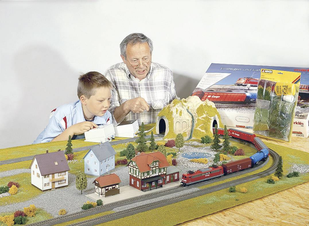 Een oudere man en een jongen bouwen samen een modelspoorbaan met miniatuurhuisjes, bomen en een tunnel op een tafel.