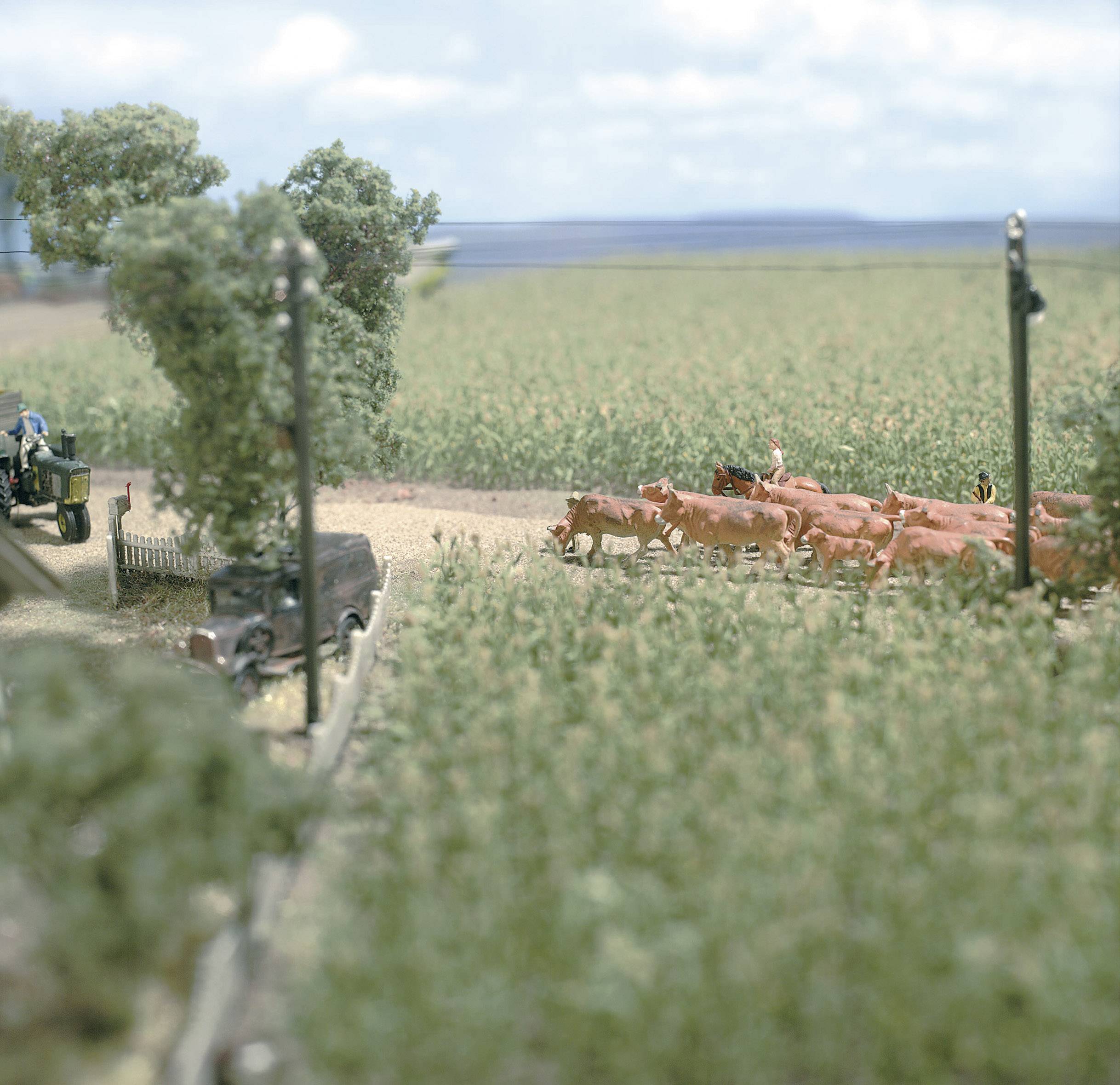 Een miniatuurscène toont een tractor op een veldweg naast een groep koeien op een weiland, omringd door bomen en veld.
