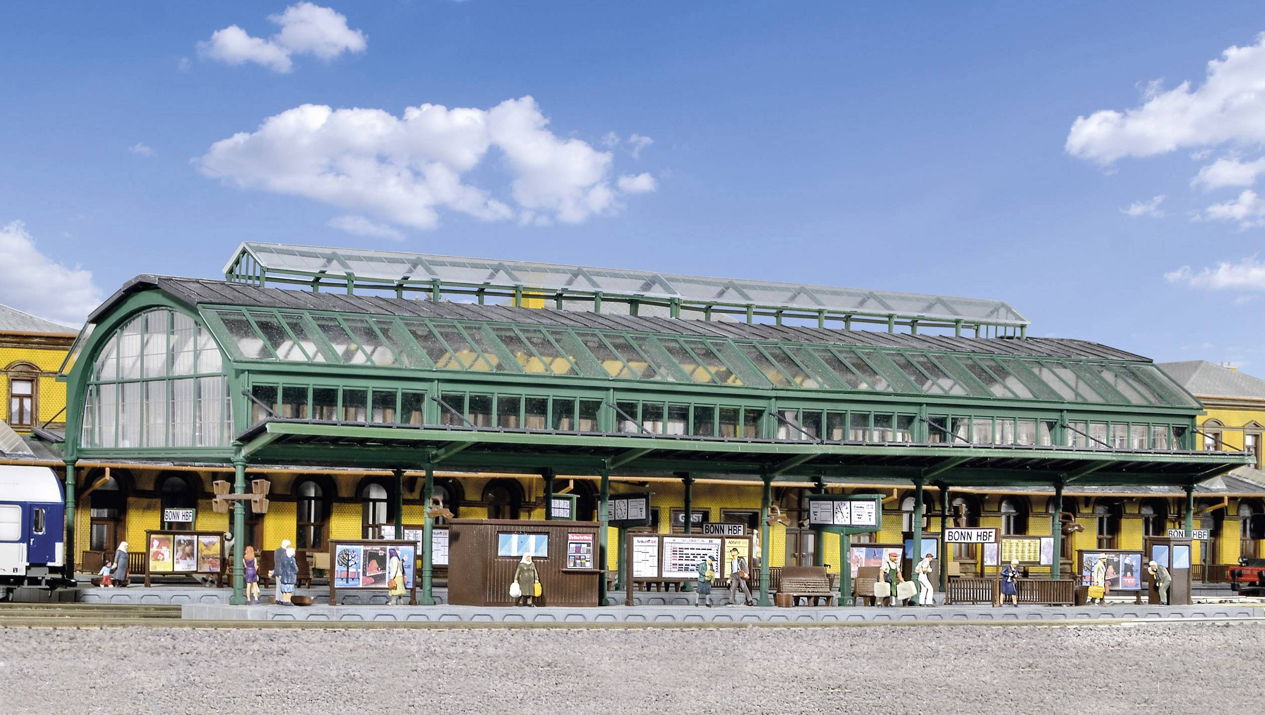 Een historisch stationsgebouw met een glazen dak en groene metalen omlijsting. Mensen staan op het perron naast reclameborden.