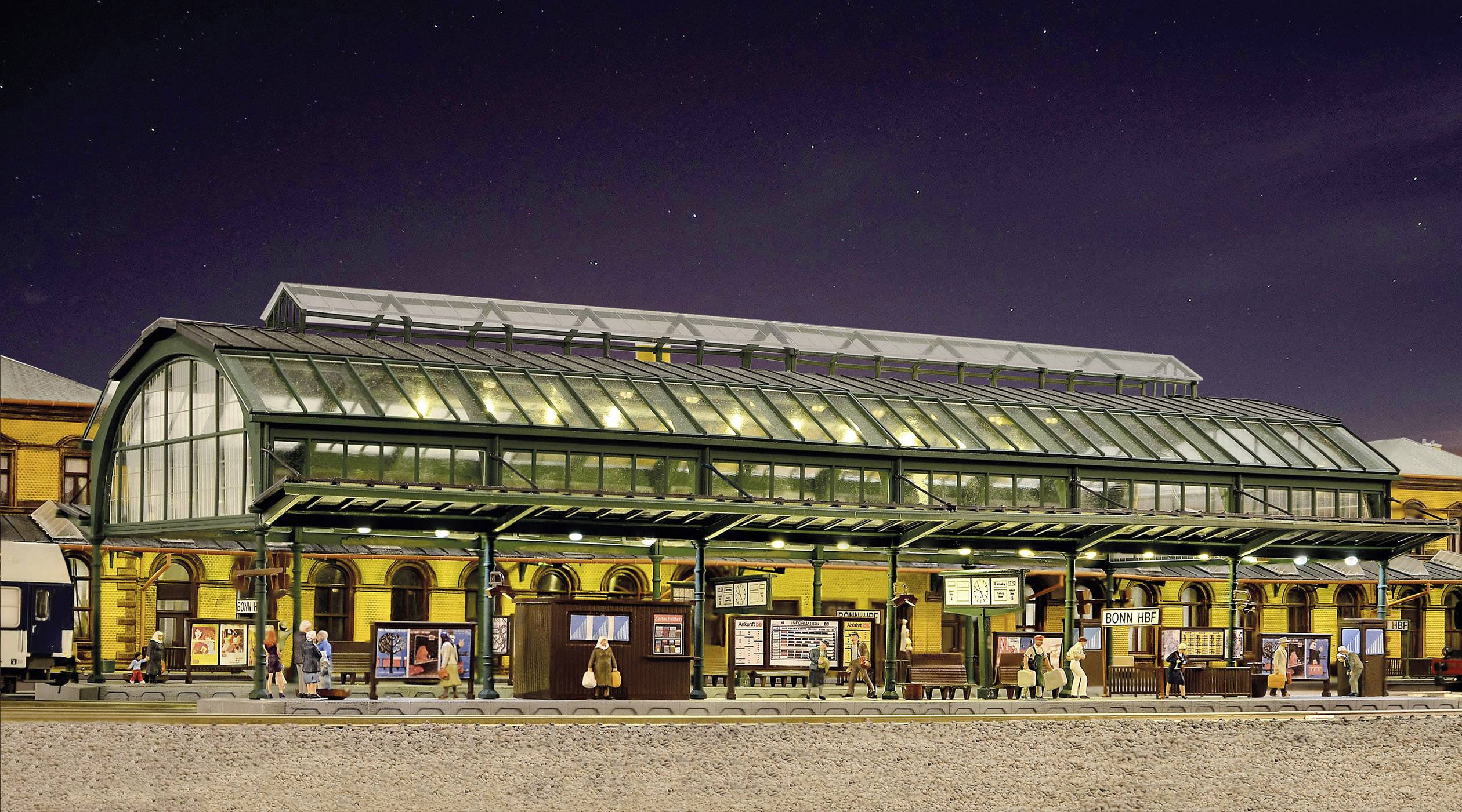 Stationshal bij nacht met historische glazen overkapping. Verschillende personen staan op het perron. Gebouwen op de achtergrond verlicht.